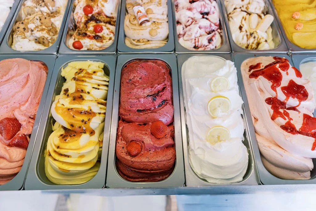 Various flavors of gelato in metal trays, including strawberry, banana with caramel, berry, lemon, and strawberry with strawberry syrup.