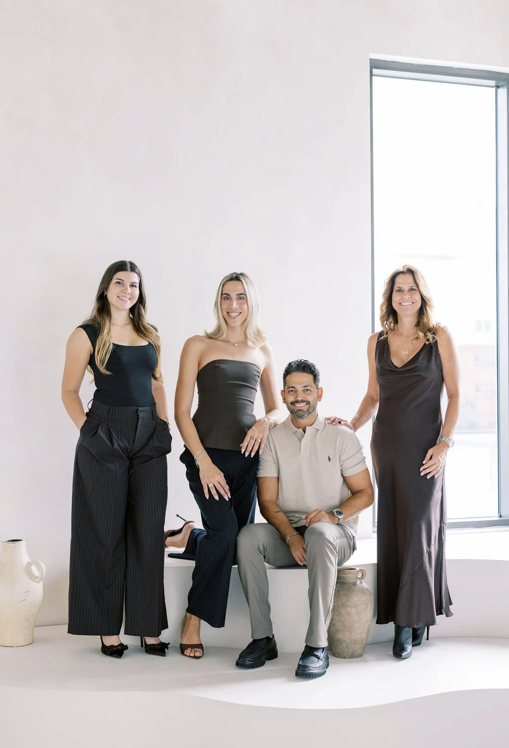 Group of four diverse professionals, three women and one man, posing indoors near a large window with natural light, smiling at the camera.