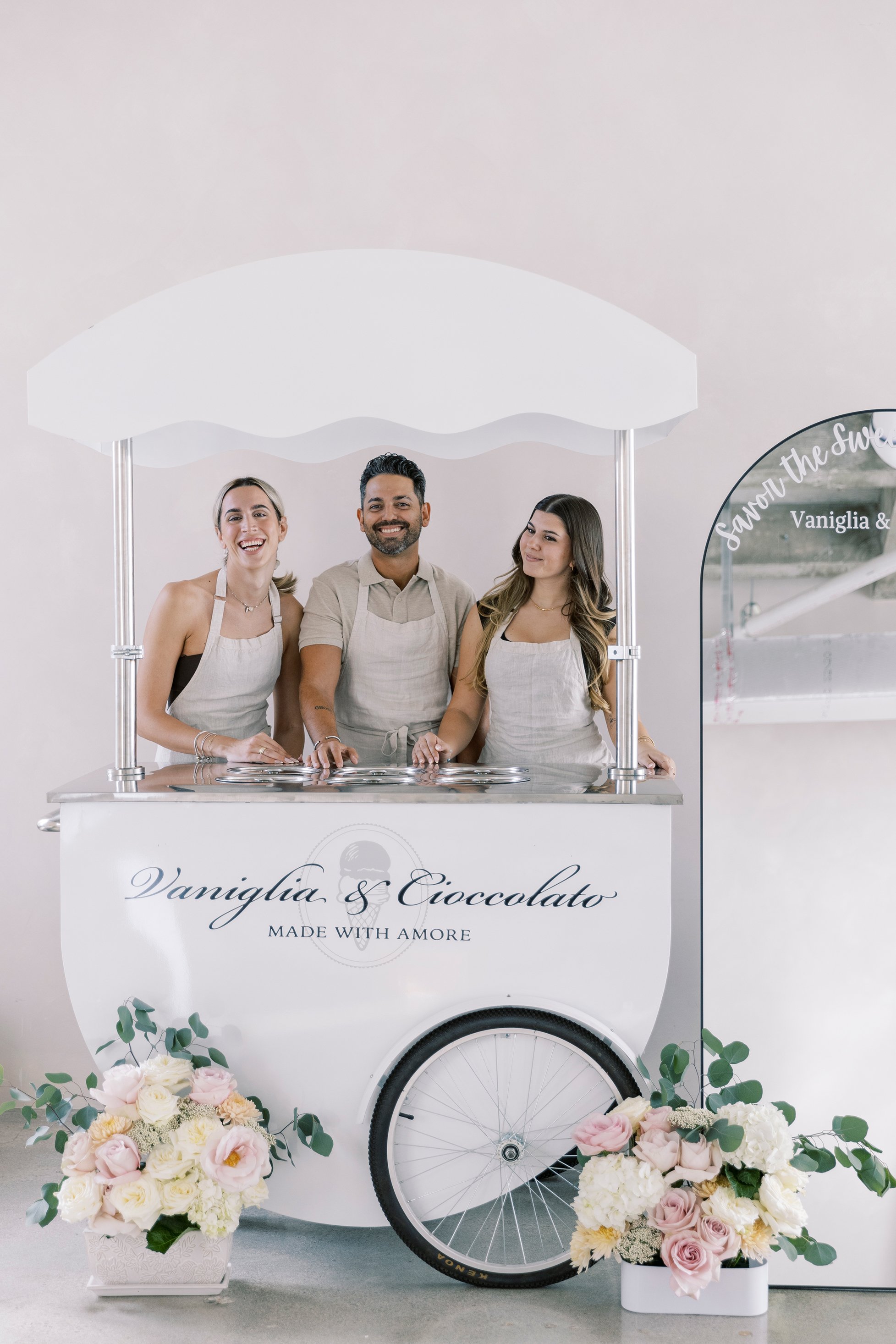 Three people standing inside a white ice cream cart with the words 'Vaniglia & Cioccolato Made with Amore' on the front, smiling at the camera, surrounded by floral arrangements, with a mirror on the right side.