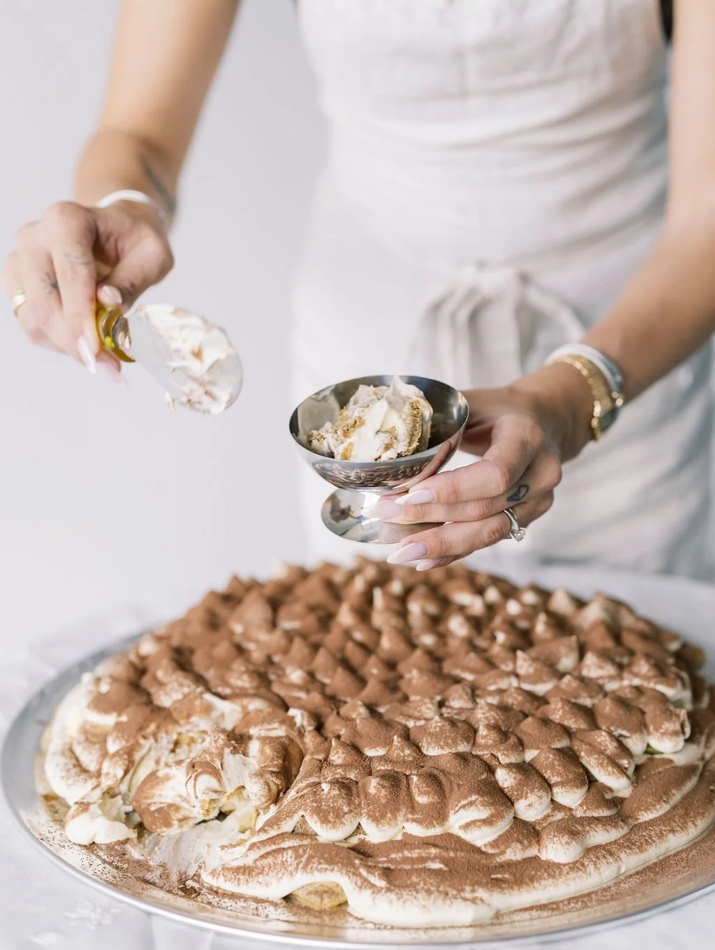 Person in white apron scooping tiramisu dessert with a spoon over a large, round tiramisu cake topped with cocoa powder and cream.