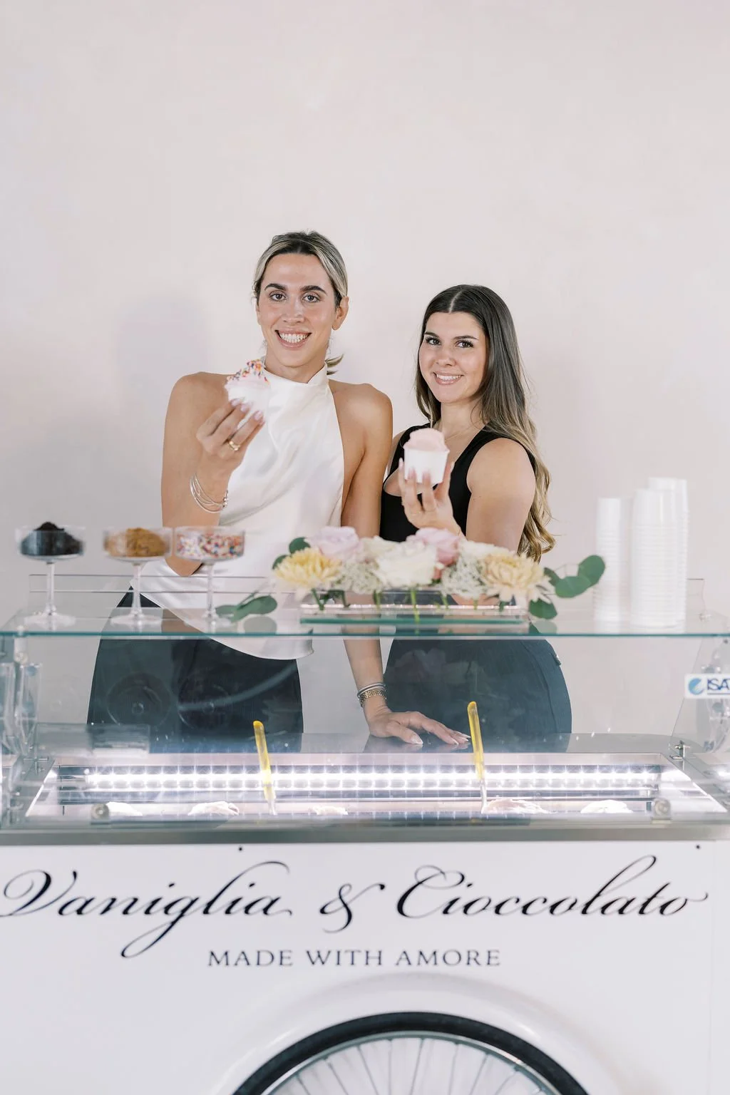 Two women with long hair smiling behind a gelato cart, holding cupcakes, with flowers on top, and stacks of cups nearby.