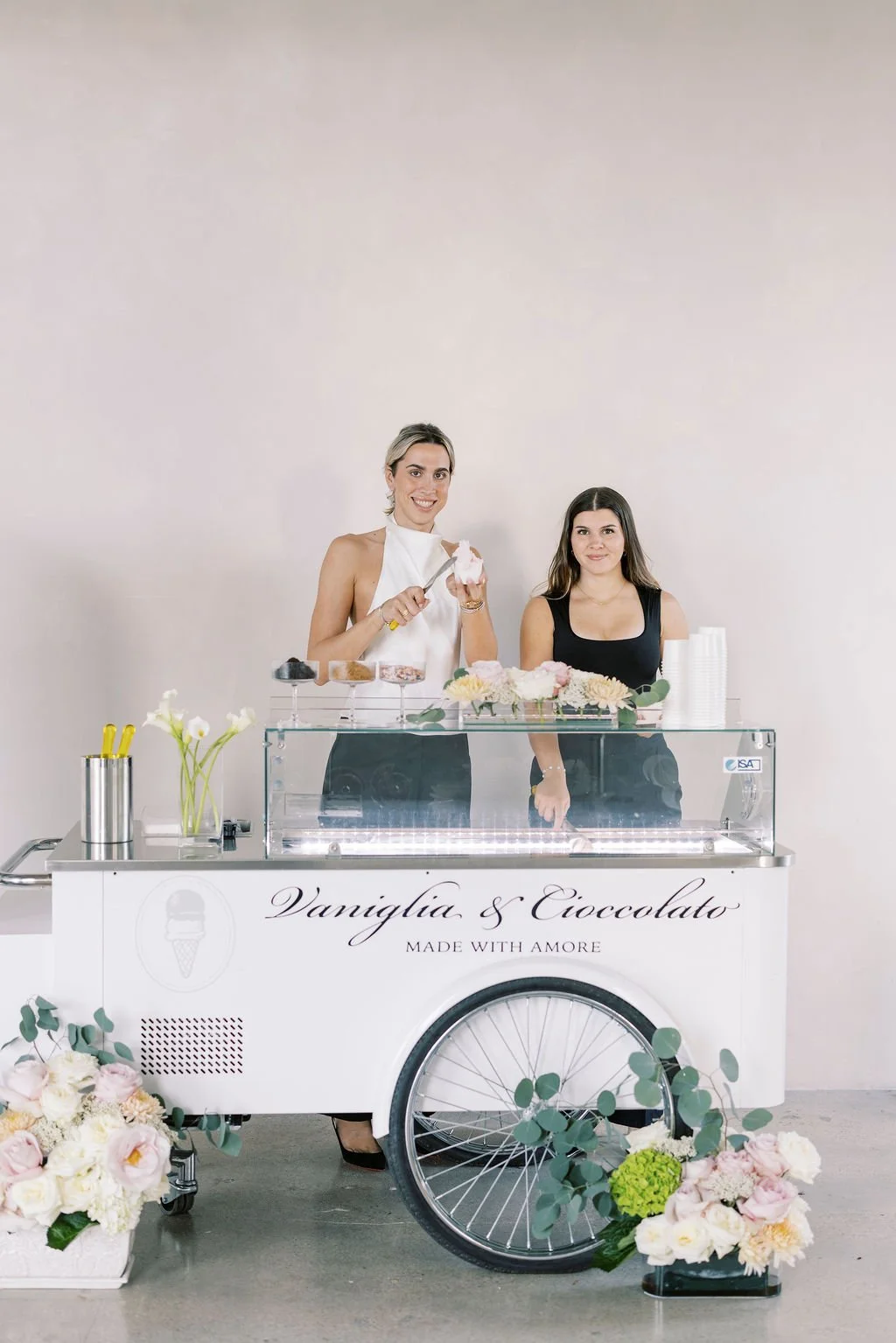 Two women standing behind an ice cream cart decorated with flowers and greenery, with one woman serving ice cream and smiling, and the other woman standing beside her. The cart has the text "V'aniglia & Cioccolato" and "Made with Amore" on it, along with a logo of an ice cream cone.