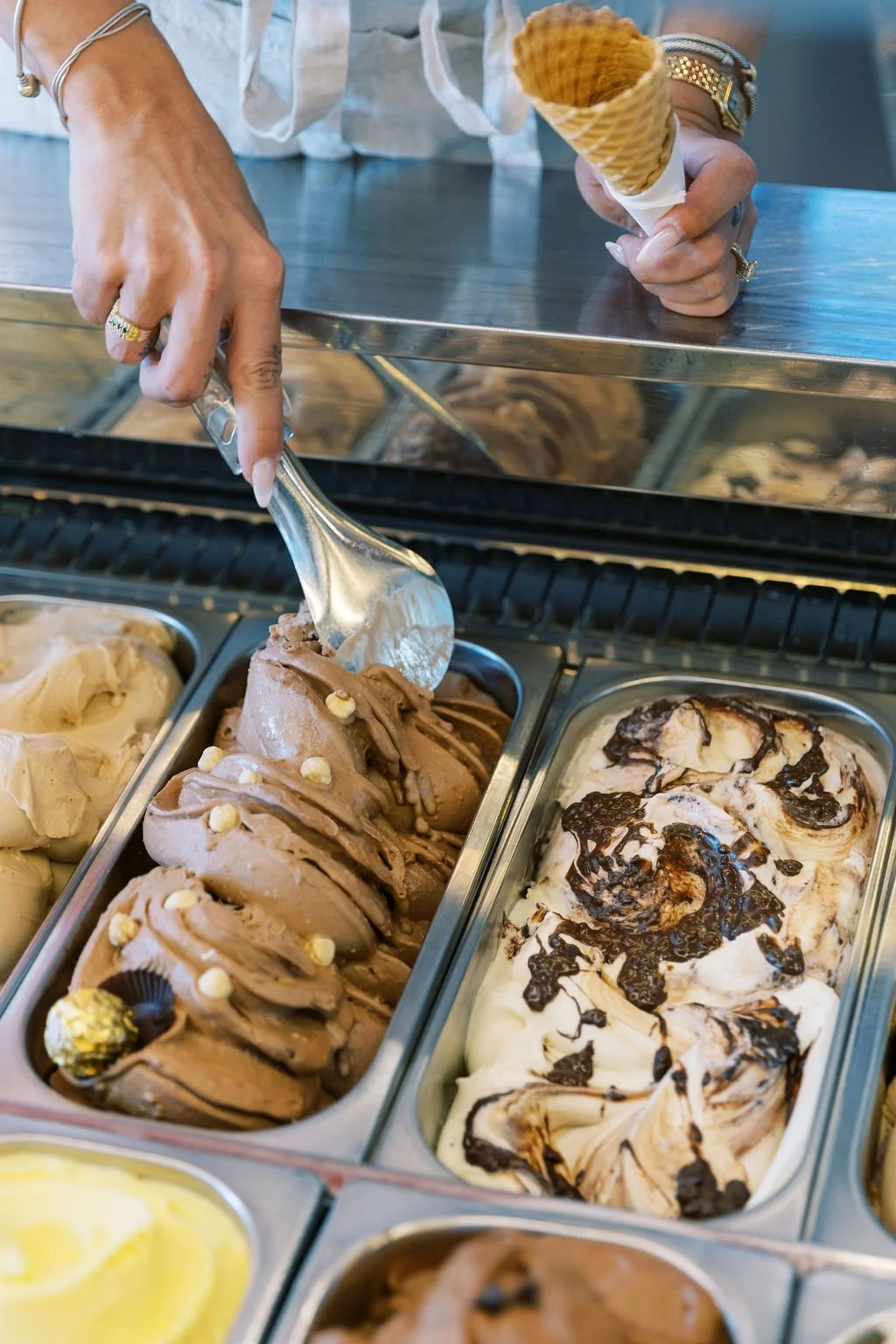 Person scooping chocolate ice cream from a metal container into an ice cream cone at a store, with other ice cream flavors visible nearby.
