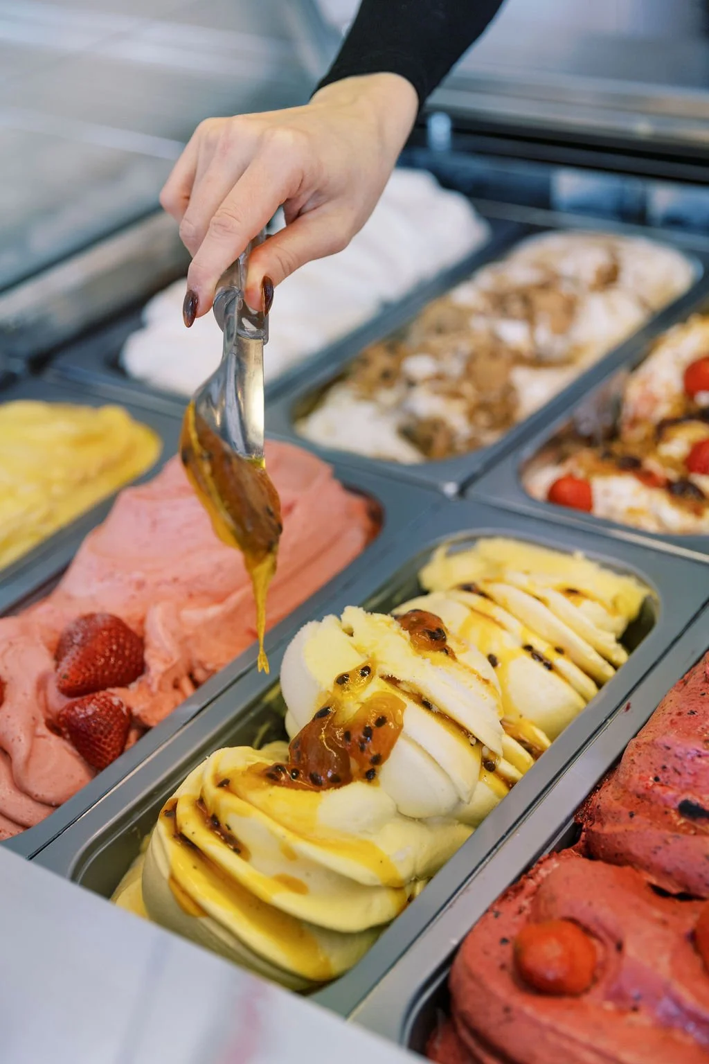 A person serving themselves vanilla ice cream topped with caramel sauce from a self-serve ice cream counter, with various other flavors of ice cream in the background.