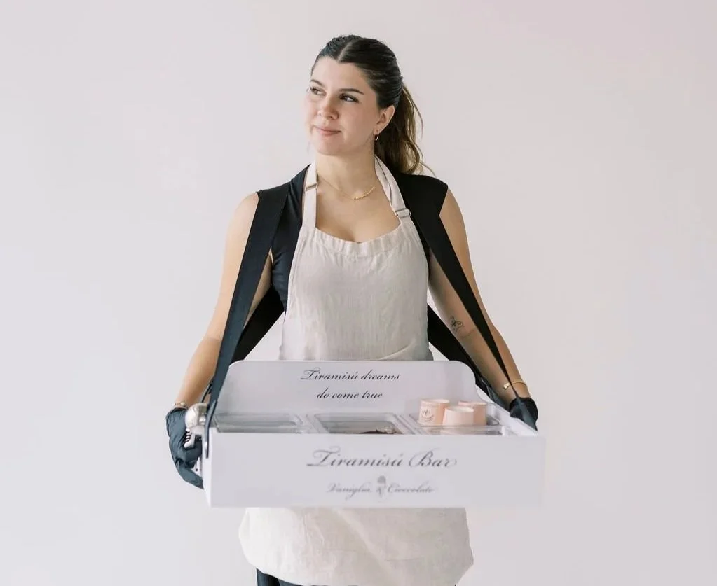 A woman wearing a black apron and gloves holding a tray with small containers, against a plain light background.