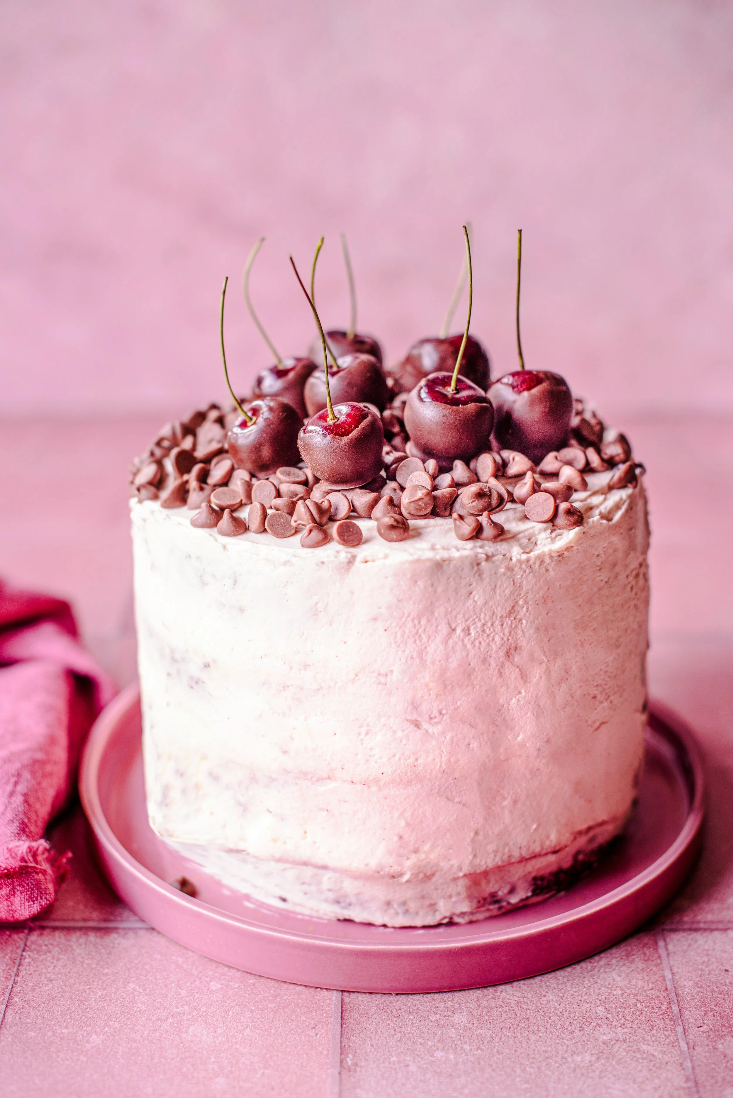 Pink frosted cake topped with cherries and chocolate chips on a pink plate.