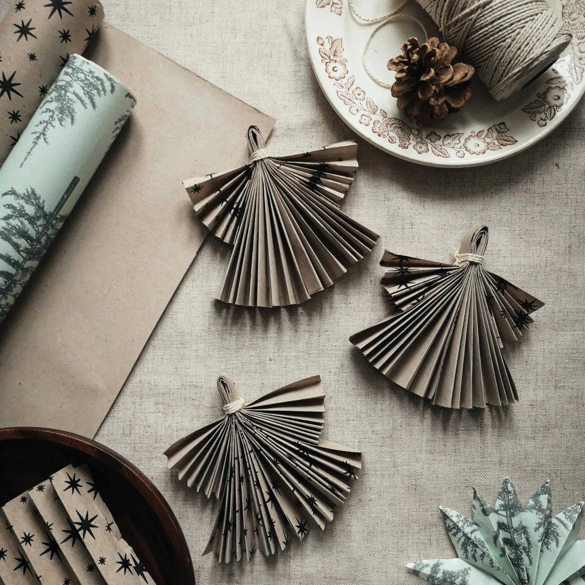 Decorative folded paper angels with star patterns, pine cone, and Christmas-themed items on a neutral fabric surface.