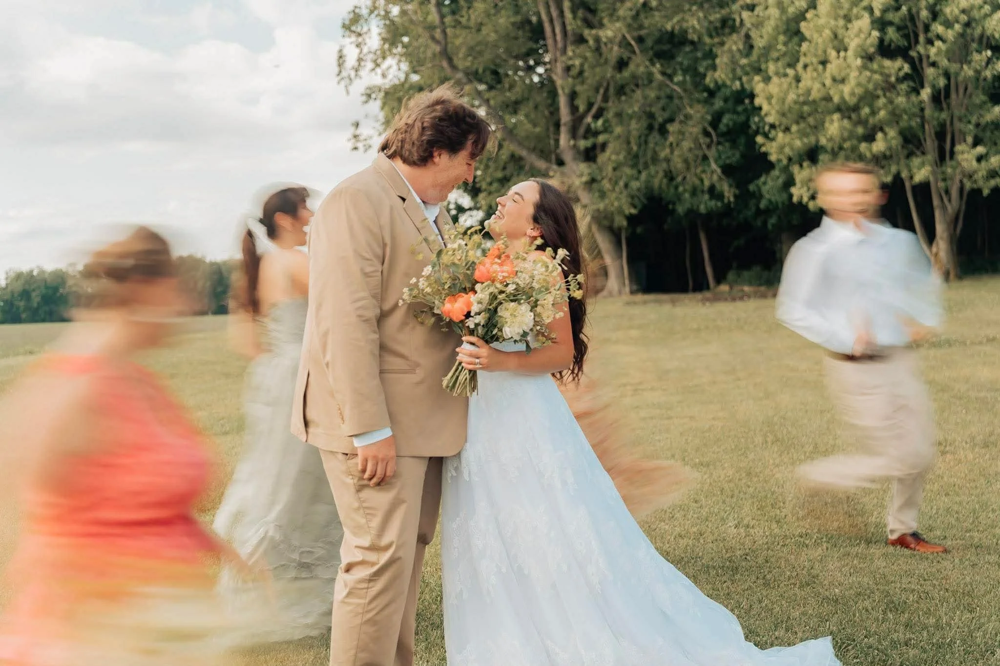 A bride and groom share a happy moment at an outdoor wedding, surrounded by friends and greenery, with a girl in a pink dress in the foreground and others blurred in the background.