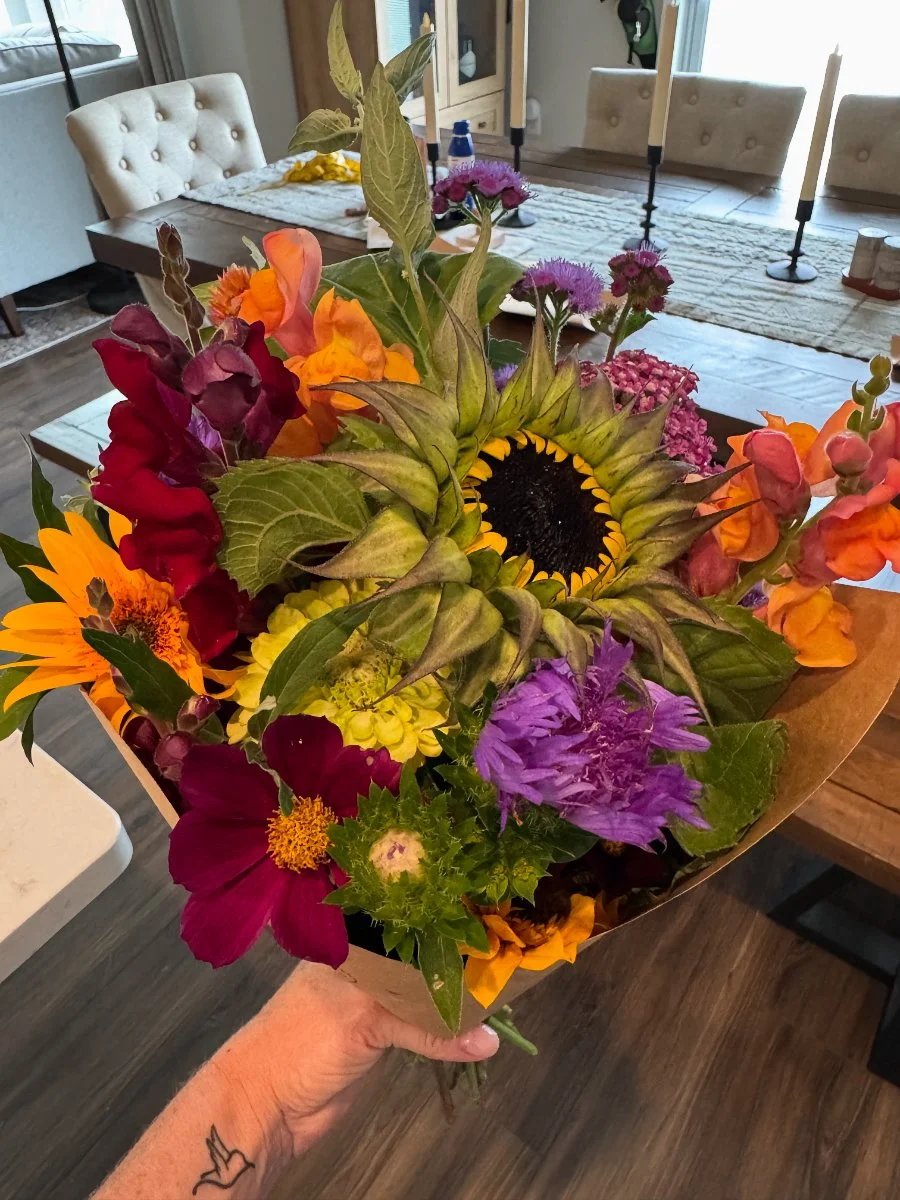 A hand holding a colorful bouquet of flowers including sunflowers, lilies, and other vibrant blooms in a living room with chairs, a table, and candles in the background.