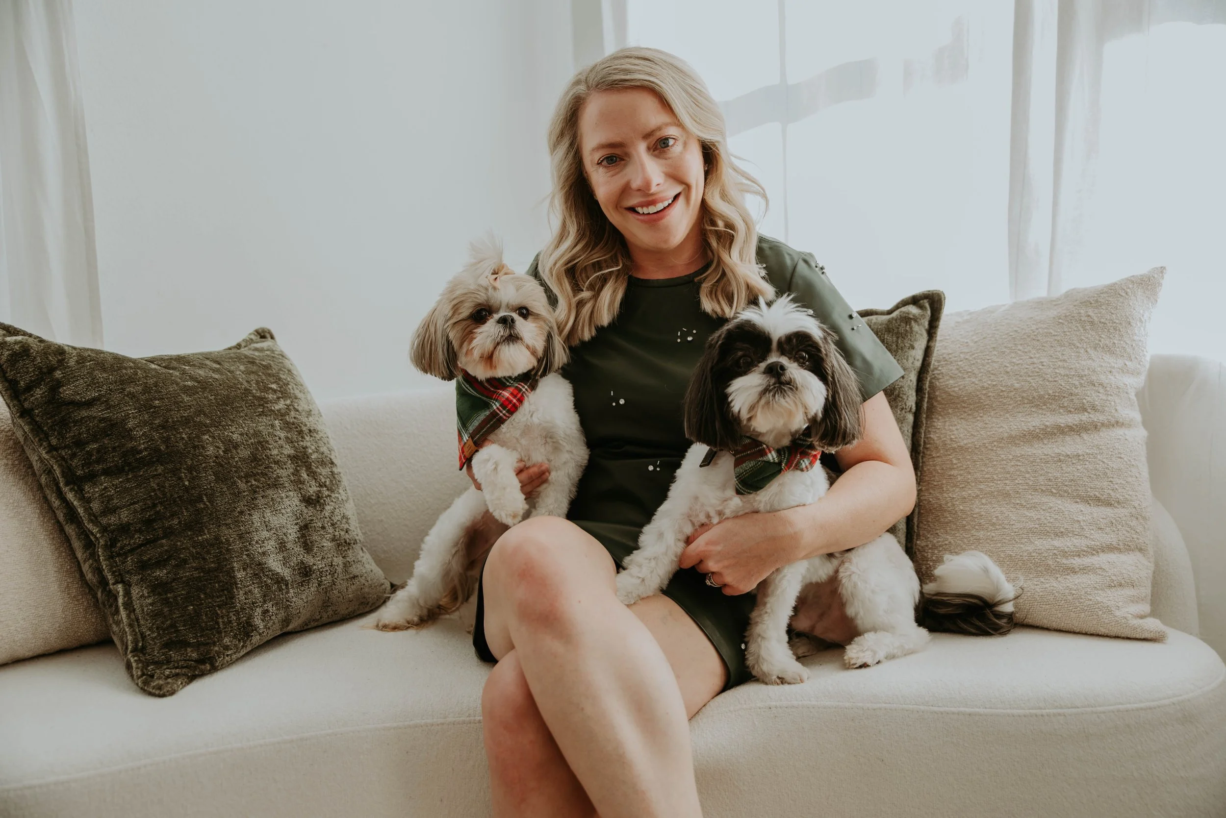 A woman sitting on a beige sofa holding two small dogs, one with a bow and the other with a bandana, both wearing Christmas scarves, in a cozy, well-lit living room.