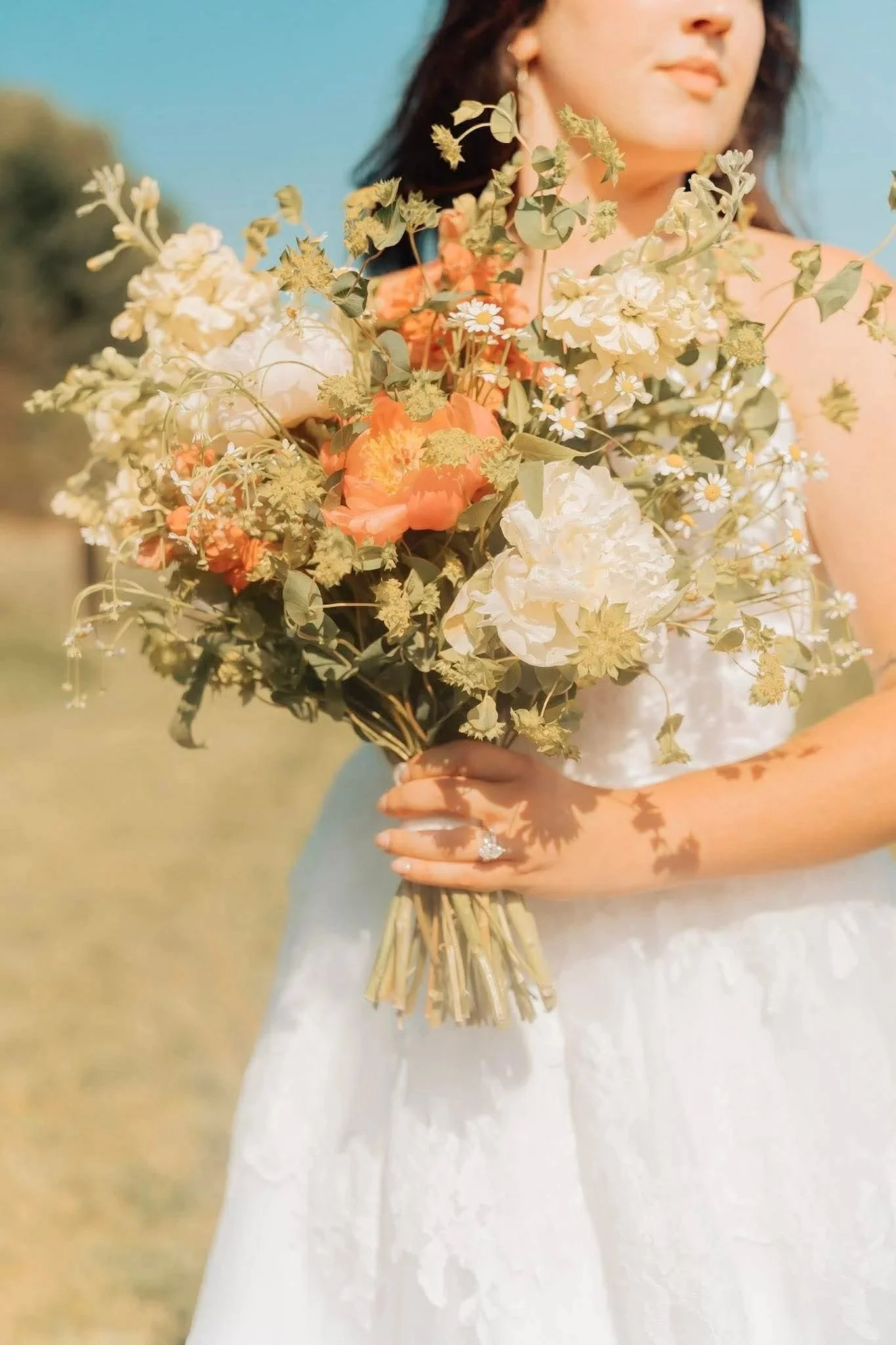 A woman in a white dress holding a large bouquet of flowers outdoors, with a blurred background and a clear blue sky.
