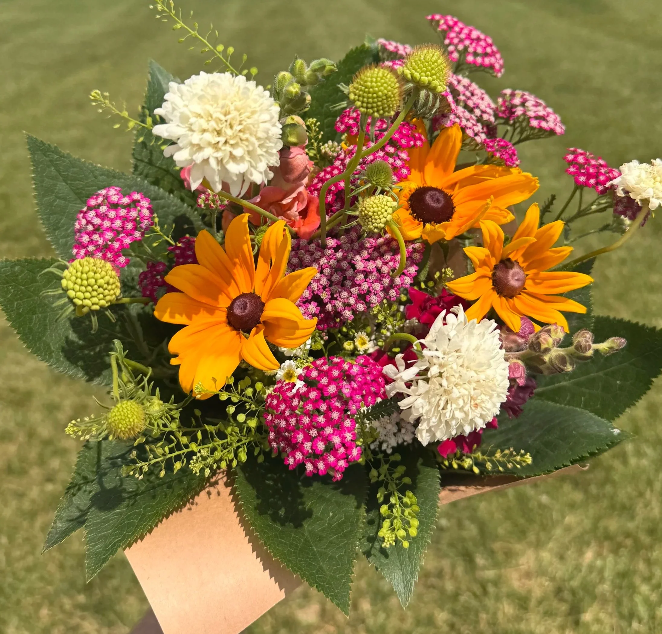 A hand holding a colorful bouquet of flowers including sunflowers, lilies, and other vibrant blooms in a living room with chairs, a table, and candles in the background.