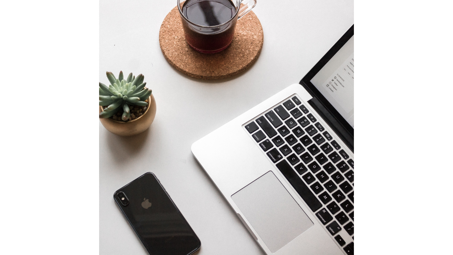 Un ordinateur portable sur une table blanche, à côté d'une tasse de café sur un dessous de verre en liège, un téléphone noir et une petite plante succulente en pot.