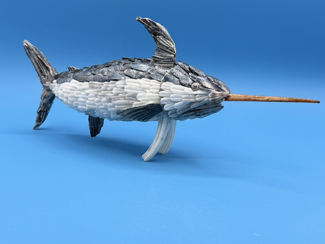 Side view of a handcrafted narwhal sculpture made from layered gray and white seashells on a blue background.