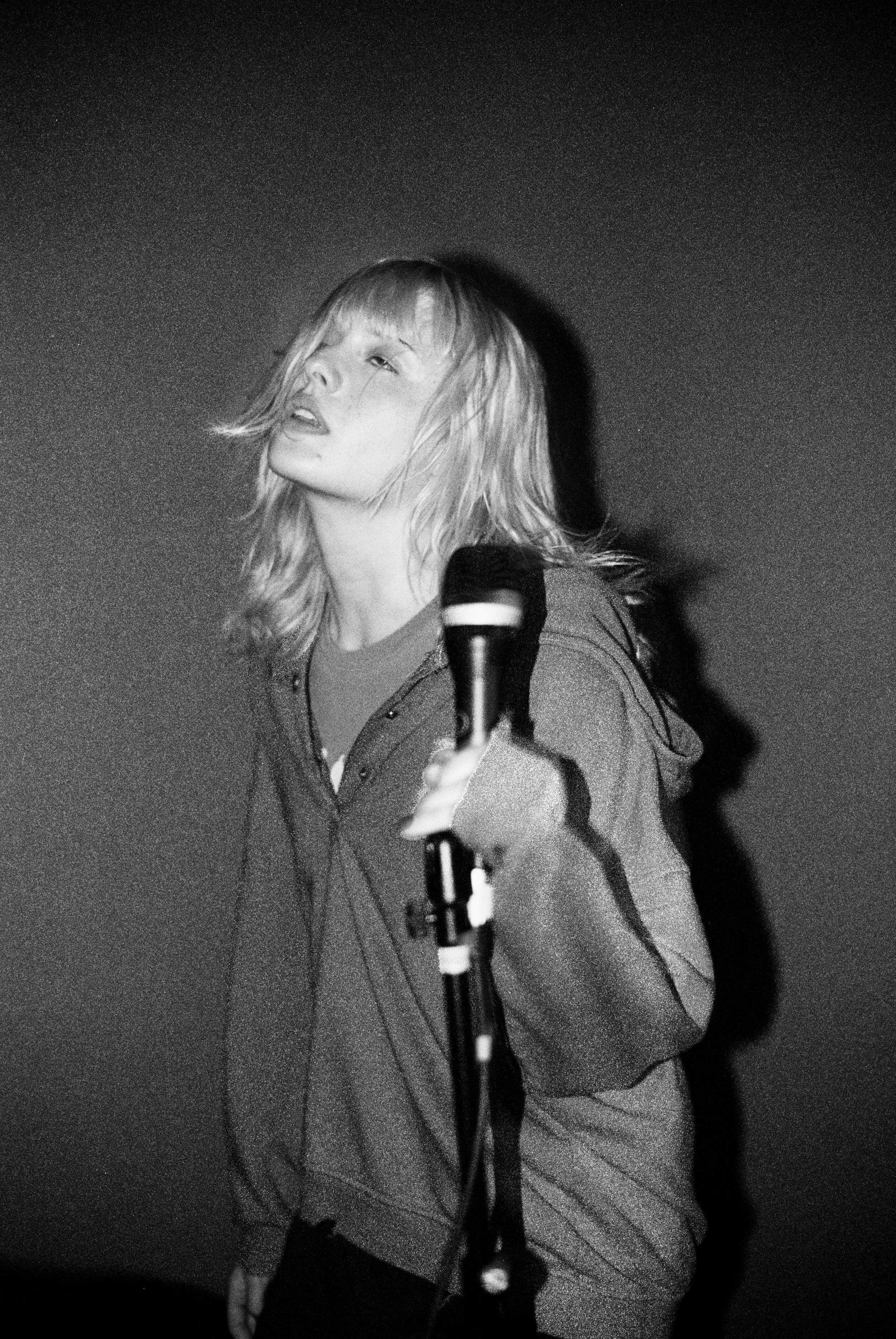 A black and white photograph of a woman with shoulder-length hair, holding a microphone, with an intense expression, in a dimly lit setting.