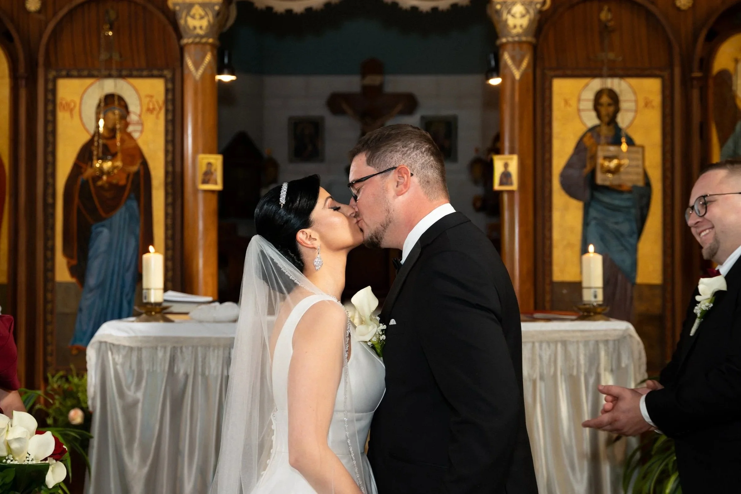 A bride and groom kiss during their wedding ceremony in a church, with religious icons and candles in the background. Image of a wedding photographed by papaleo productions.