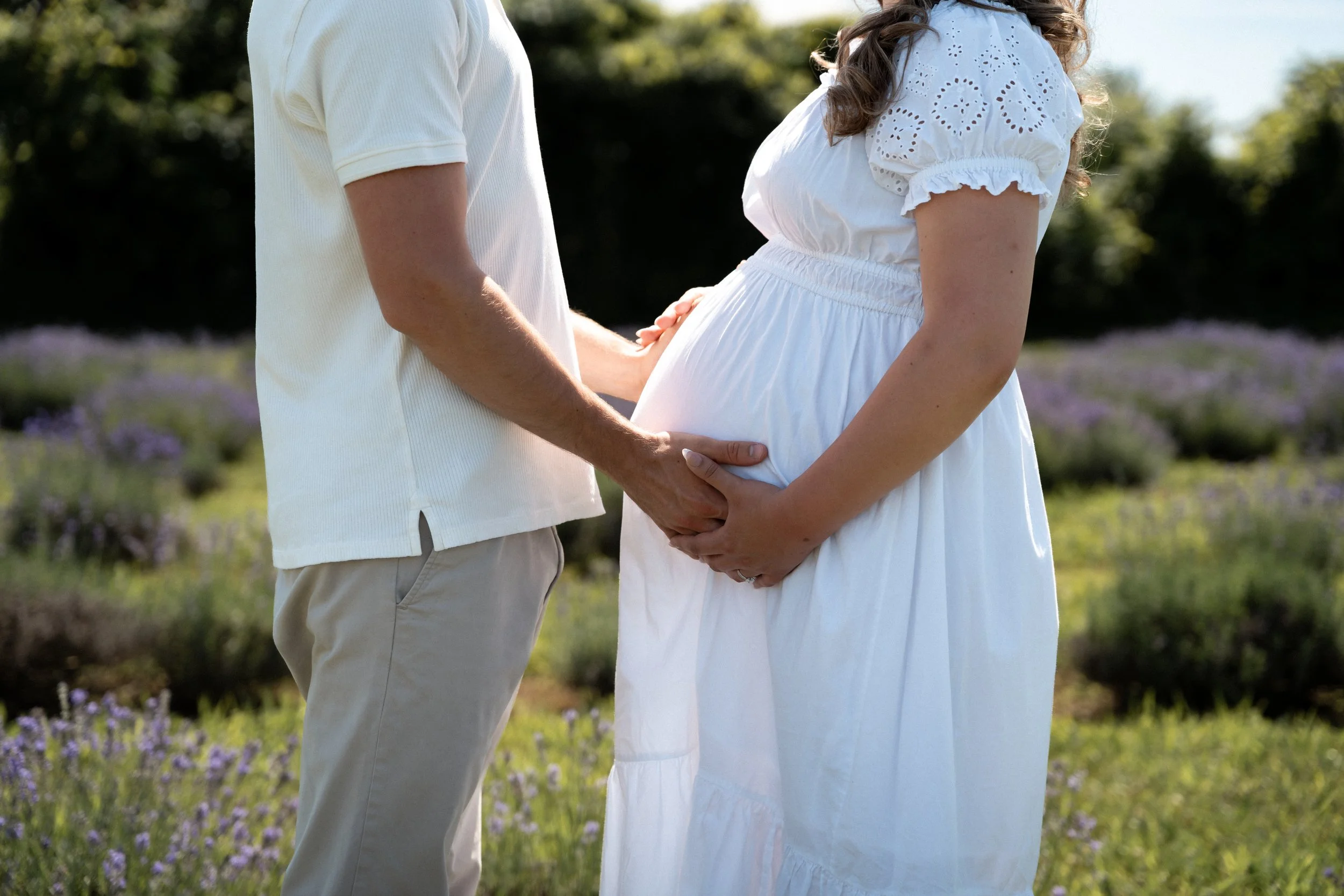 A pregnant woman and a man hold hands and touch her belly in a sunlit outdoor setting, surrounded by greenery and purple flowers.
Image of a maternity photoshoot session done by papaleo productions.