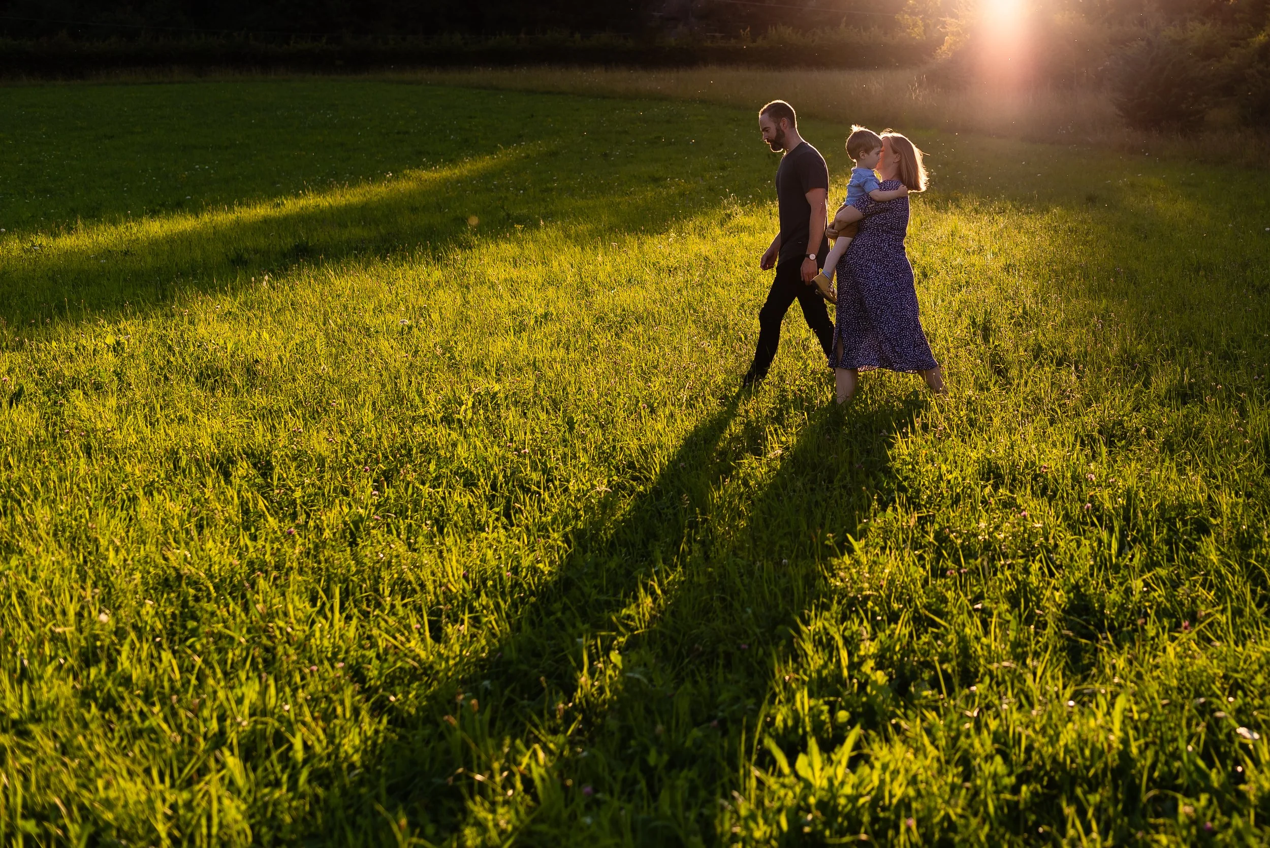 Family walking together in a grassy field at sunset, with a woman carrying a young child and a man walking nearby, captured during an outdoor family session in the botanical gardens in Geneva, Switzerland