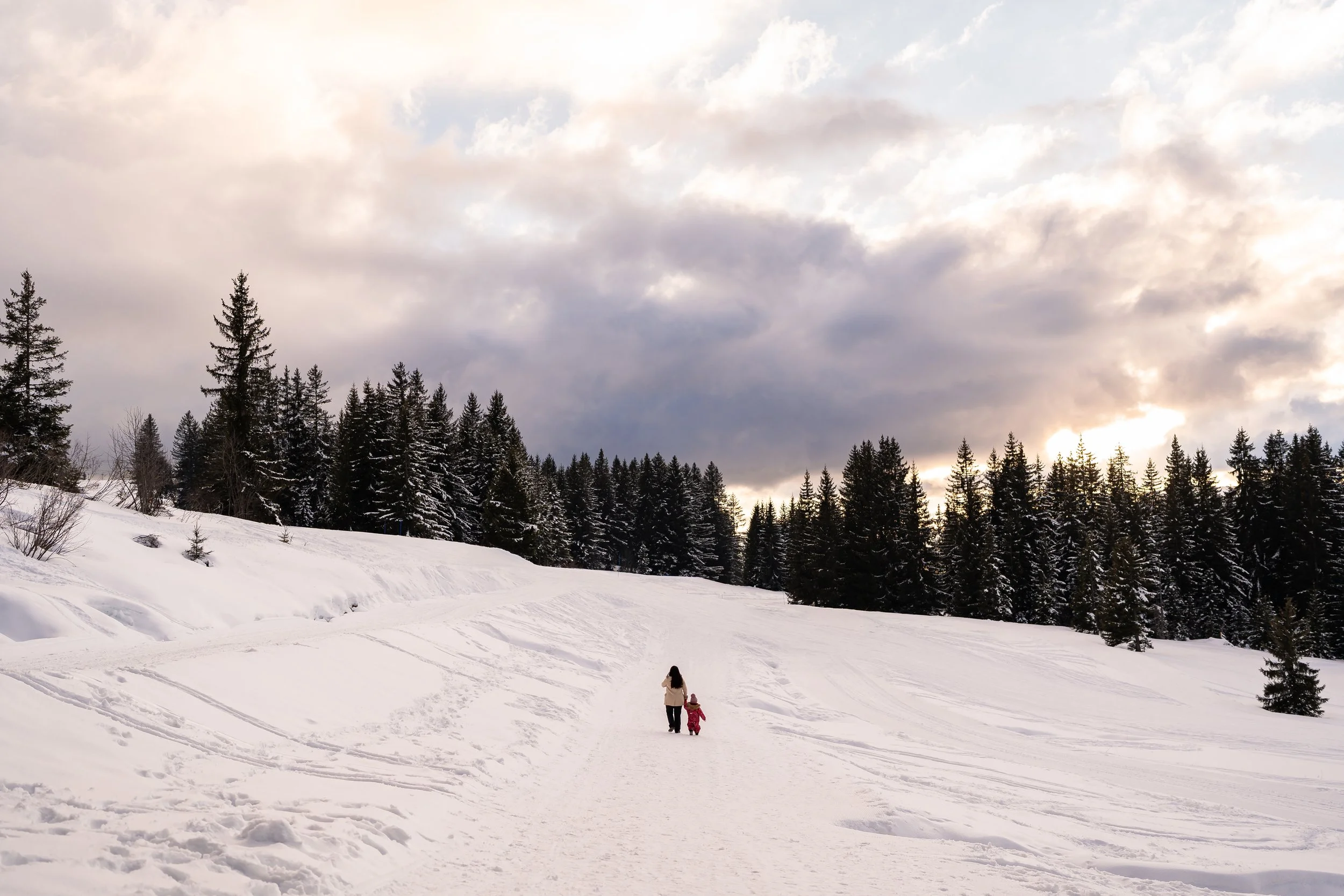 Woman and child walking on a snow-covered trail in a winter forest with tall pine trees under a cloudy sunset sky, captured during an outdoor winter family session in Les Gets in the French Alps
