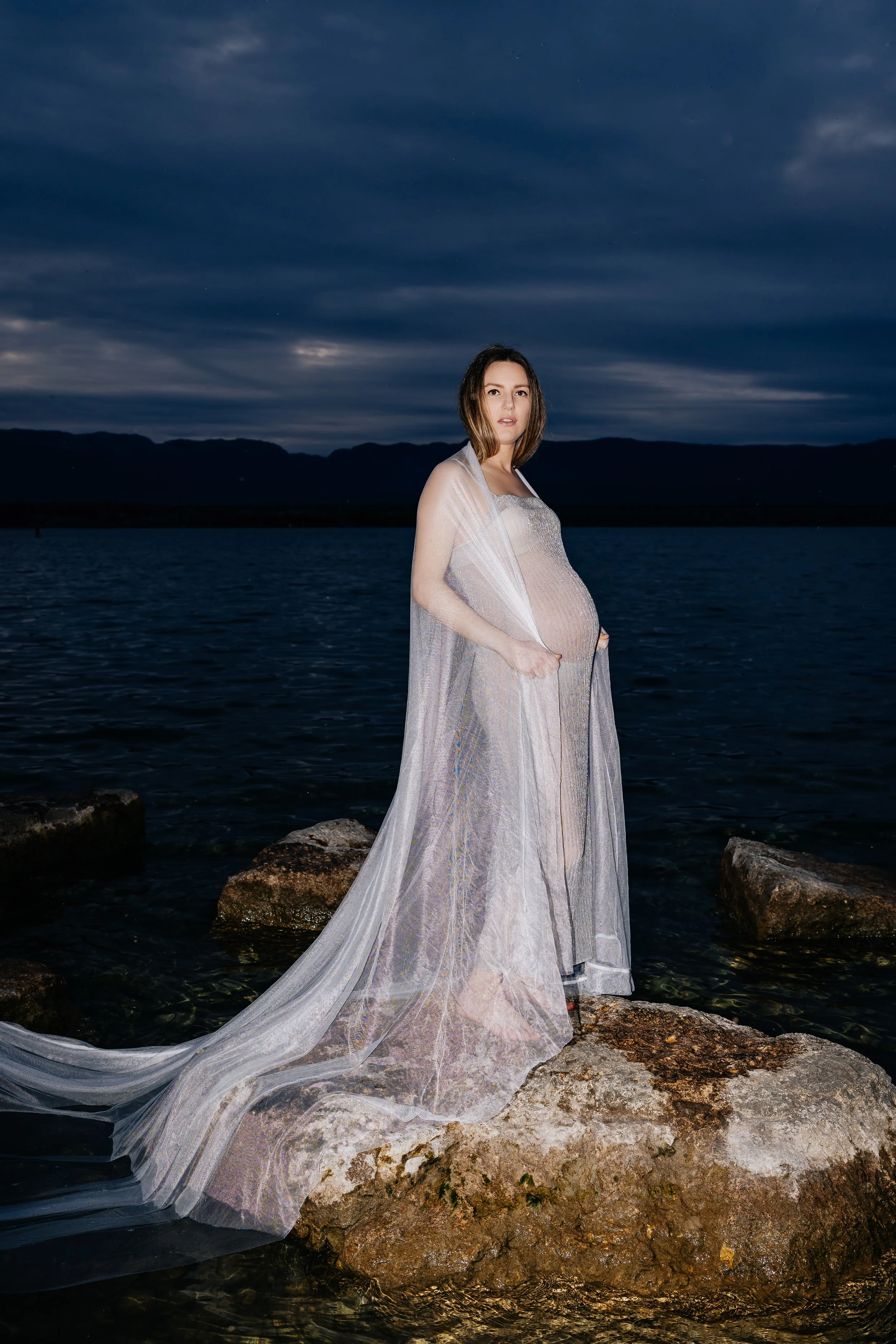 A pregnant woman standing on rocks near a body of water during nighttime, wearing a sheer, flowing gown with her hands in her pockets, with a dark, cloudy sky in the background. Maternity photographer Geneva, Switzerland.