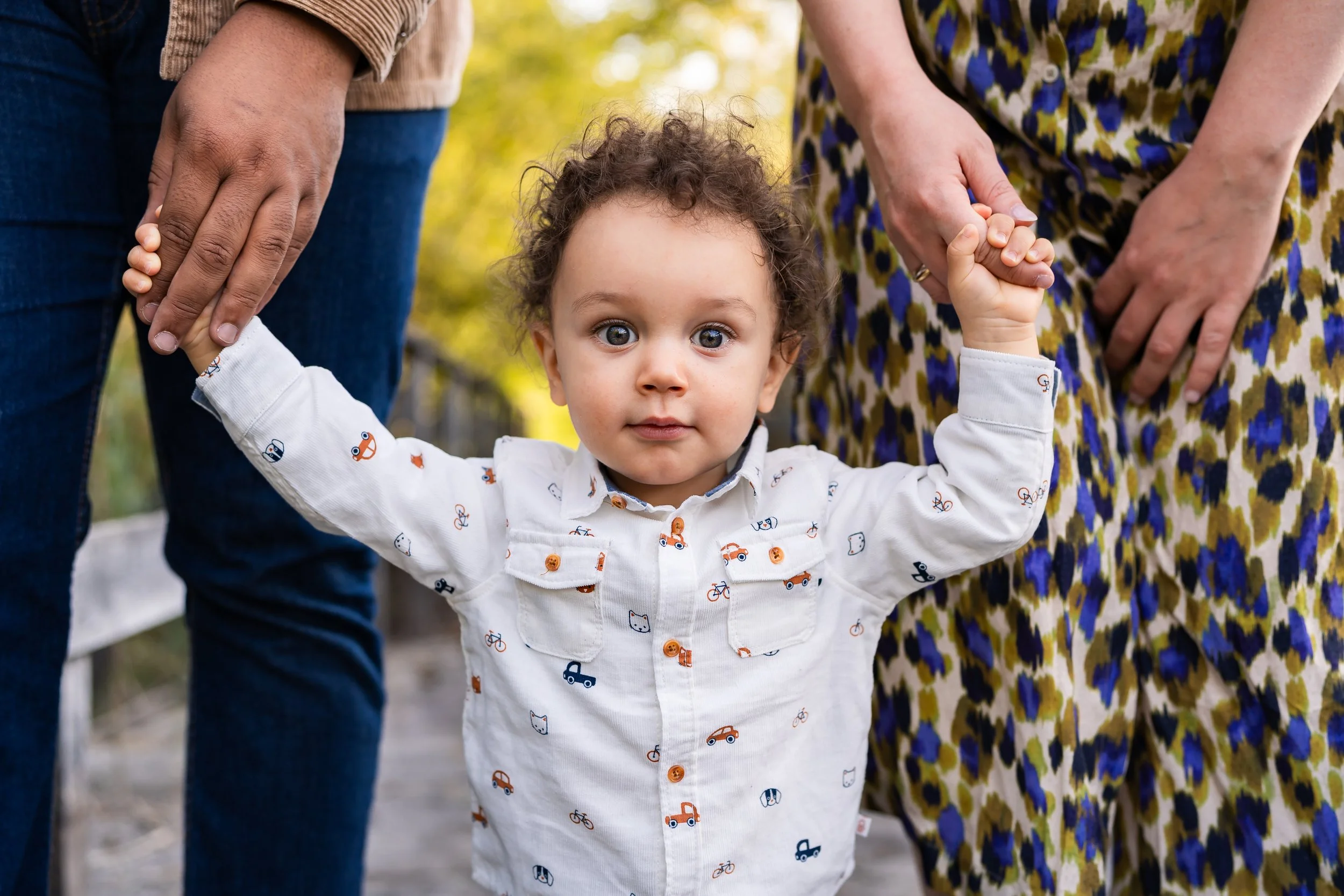 Young child with curly hair and large eyes holding hands with two adults, one on each side, outdoors during daytime, wearing a white shirt with colorful vehicle prints, captured during an outdoor family session near Geneva, Switzerland