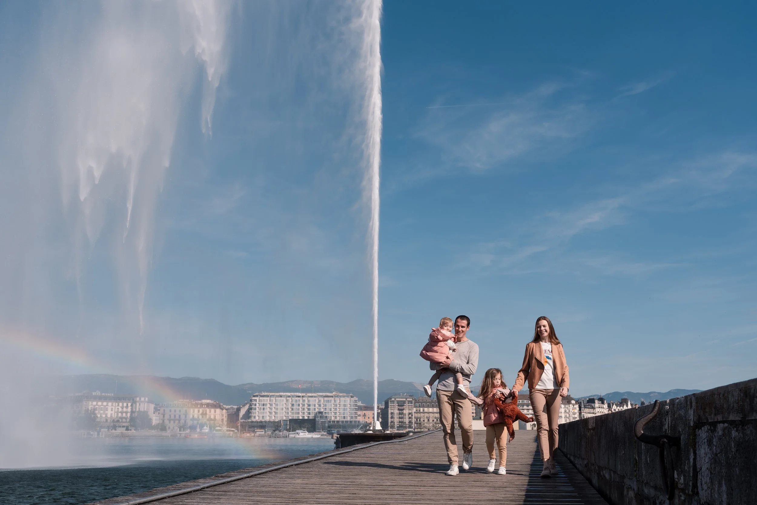 Family of four walking on a pier near the Jet d'Eau fountain, with Geneva’s cityscape in the background under a blue sky, captured during an outdoor family photoshoot in Switzerland