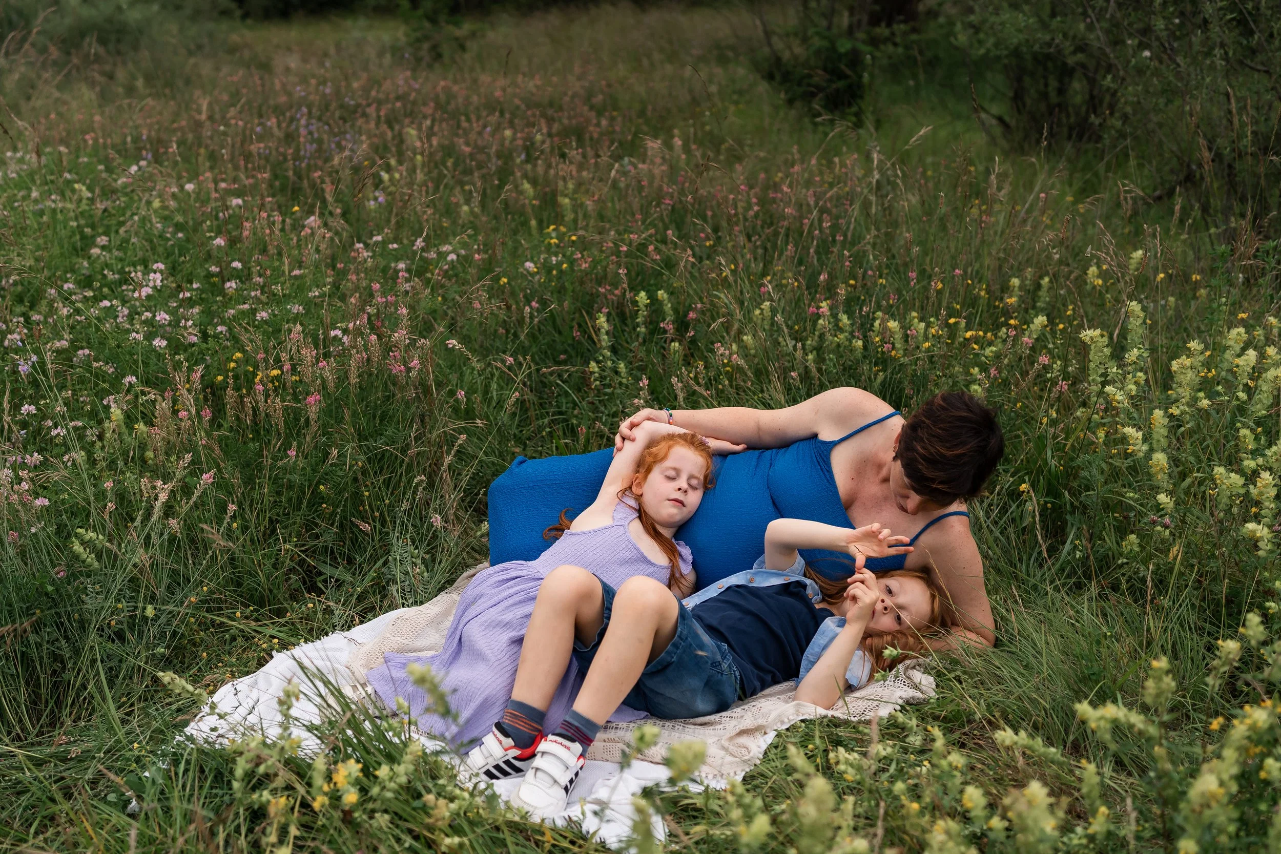 Mother lying on a blanket in a grassy field with her young son and daughter among wildflowers, relaxing together during an outdoor lifestyle family photoshoot in Geneva