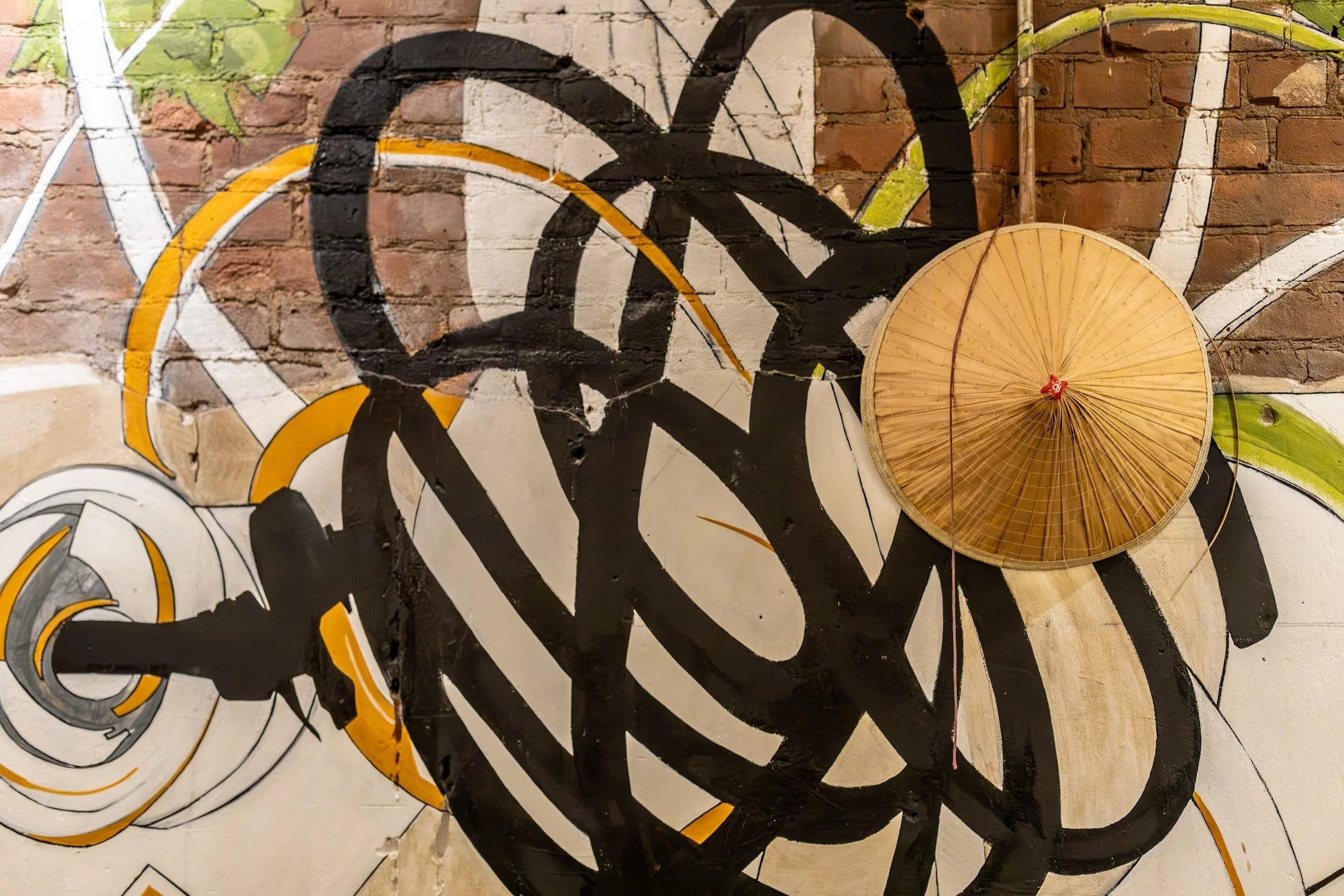 An abstract street art mural on a brick wall featuring black, white, yellow, and green swirling lines. A round, woven bamboo parasol is mounted on the wall, partially overlapping the mural at here and now brewing company in honesdale, pa.
