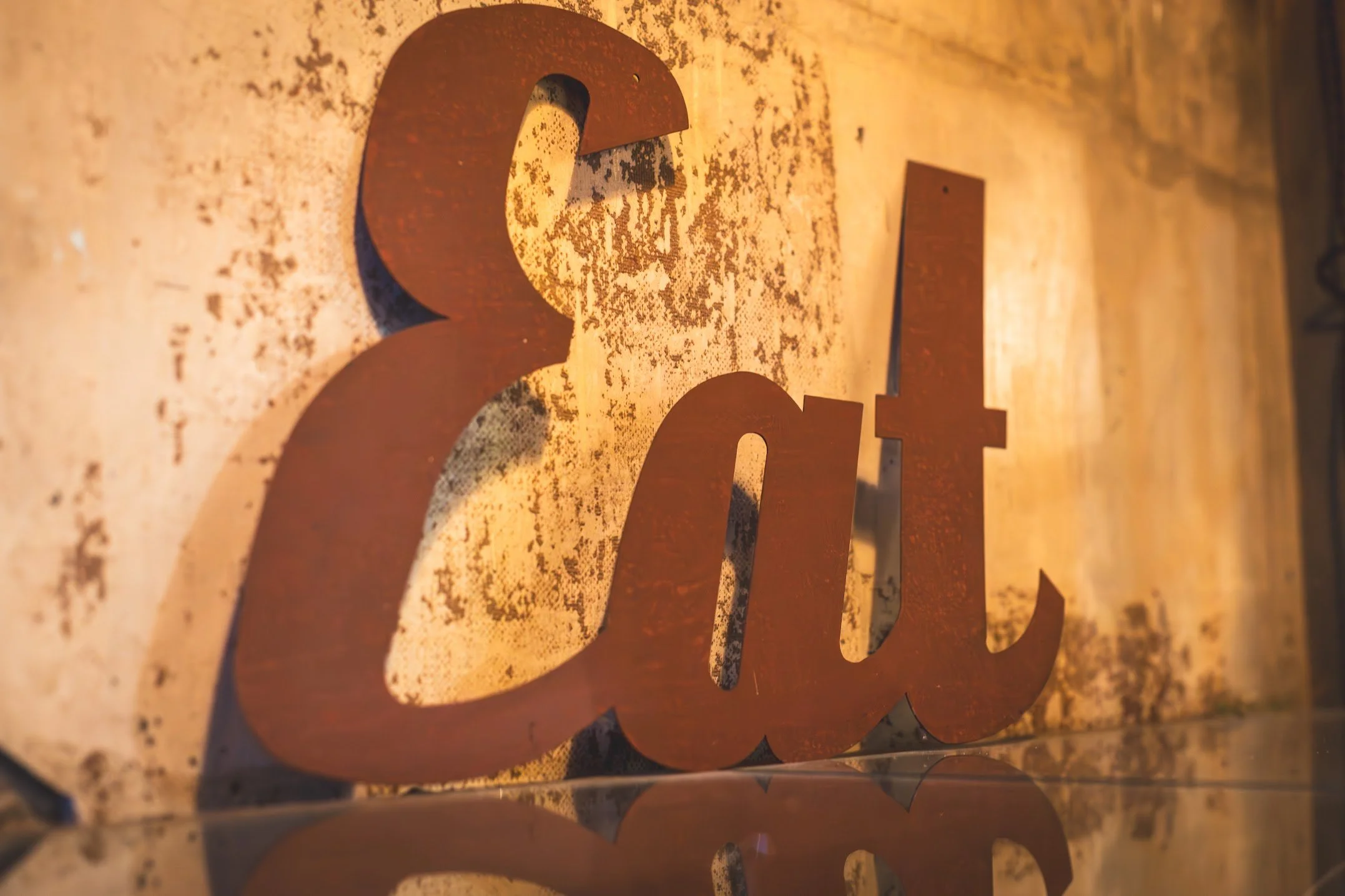 Close-up of rust-colored metallic letters spelling "eat" on a textured, distressed wall with warm lighting reflected on the surface.