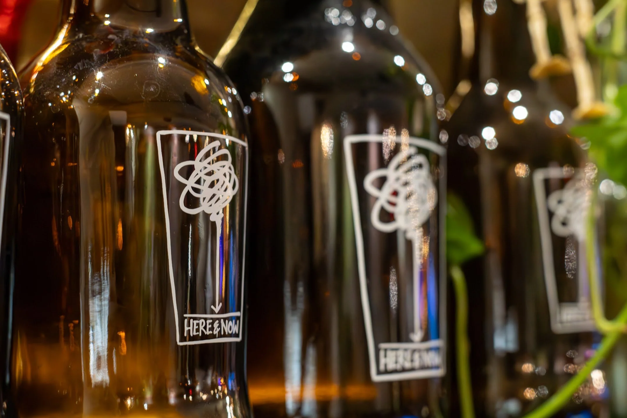 Close-up of several dark glass bottles (growlers) with white abstract designs (our logo) and the text 'HERE & NOW' on their labels, lined up with blurred background in honesdale, pa.