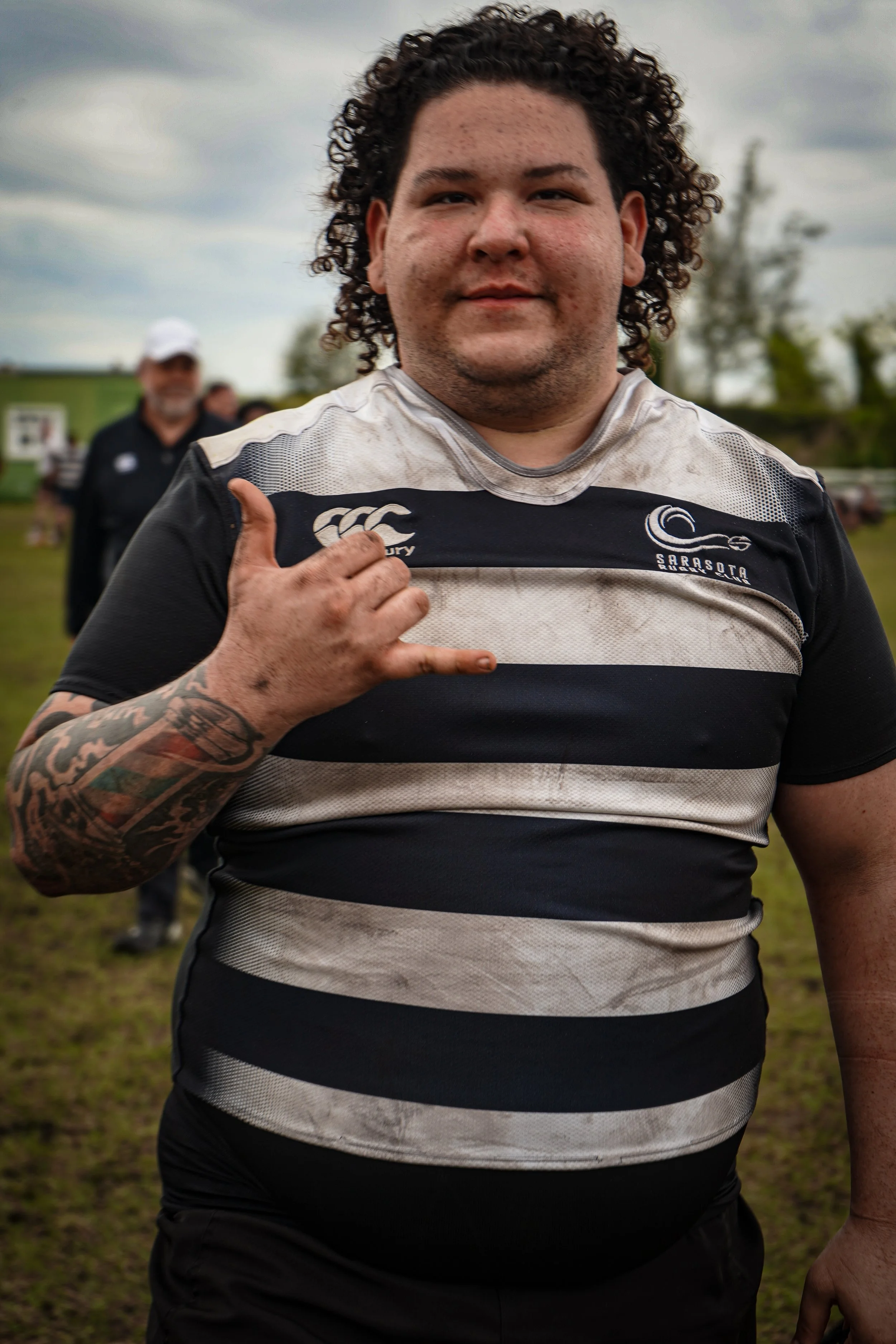 A young man with curly hair and tattoos on his left arm is standing outdoors on a cloudy day. He is wearing a black and white striped rugby shirt with a logo that says 'Sarasota, Rugby' and is making a hand gesture with his left hand. He appears to b