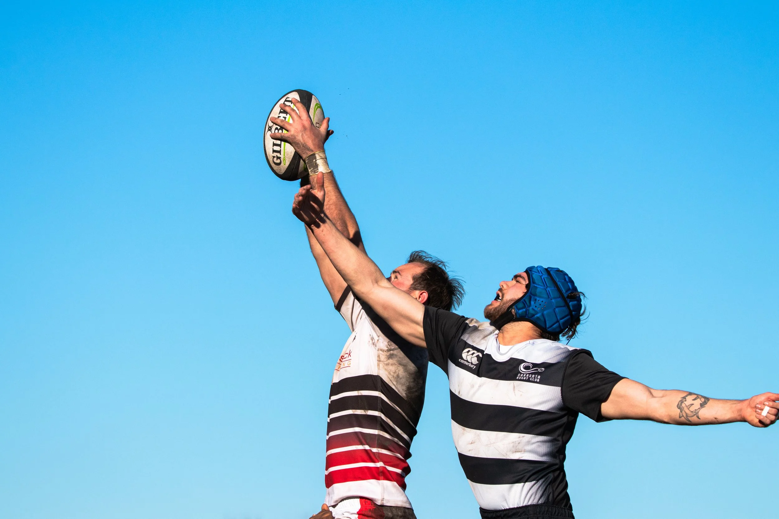 Two rugby players competing for the ball, one in a white and red uniform, the other in a black and white striped uniform with a blue helmet, against a clear blue sky.
