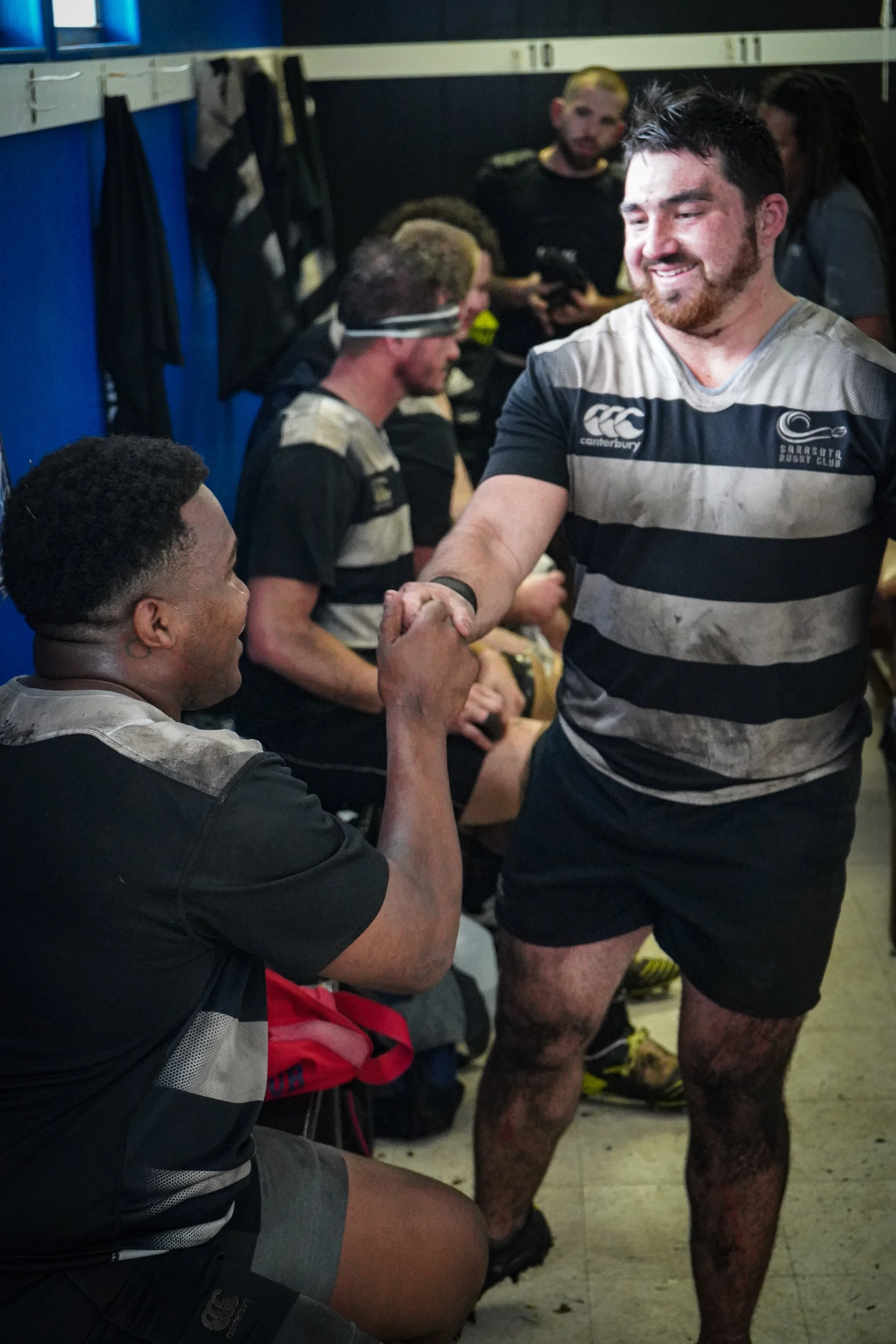 Two rugby players shaking hands in the locker room, with others sitting and standing in the background.