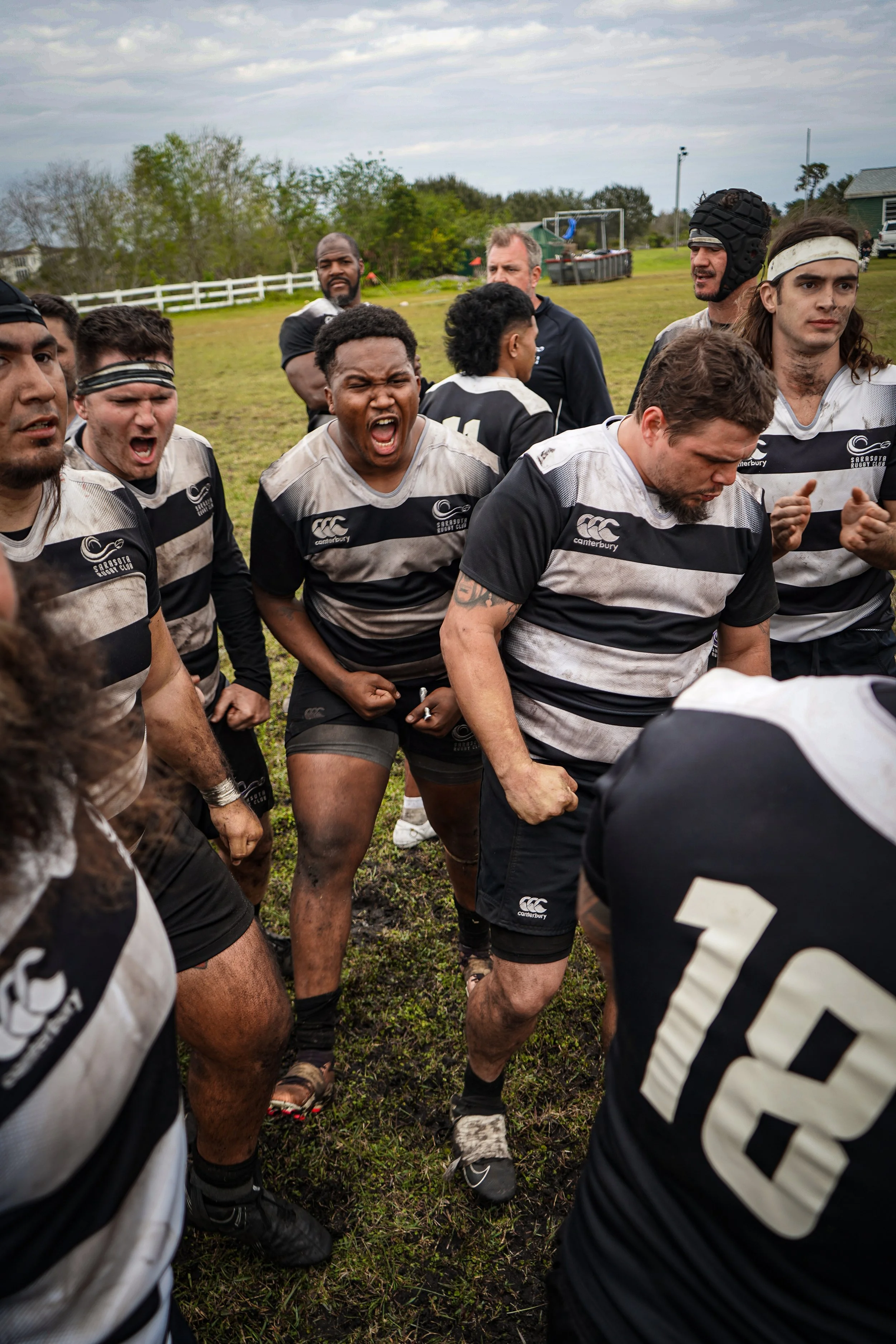 Rugby team huddled together outdoors, some players shouting and others looking focused, with muddy grass and trees in the background.