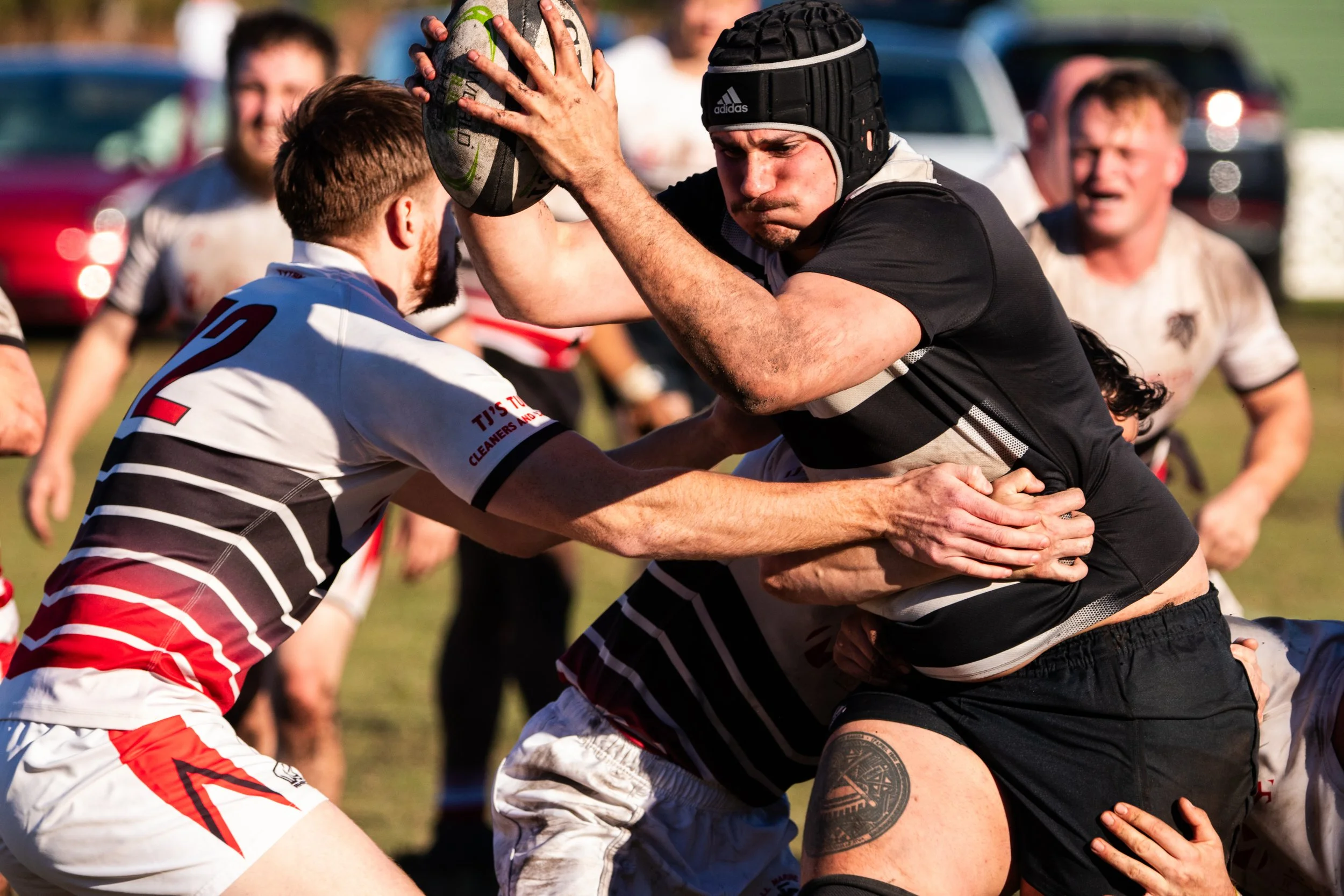 Rugby players in a tackle during a match, with players in black and white striped jerseys and others in white and red jerseys, on a grass field with vehicles in the background.