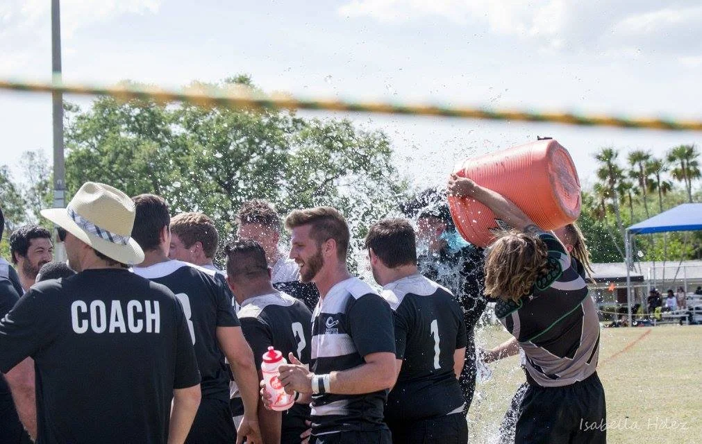 Rugby players celebrating with water splash, coach in black shirt with 'COACH' written on back, sunny outdoor field.