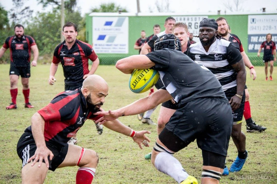 Rugby players in black and red uniforms play a tackle on a grassy field during a match, with other players and a green fence in the background.