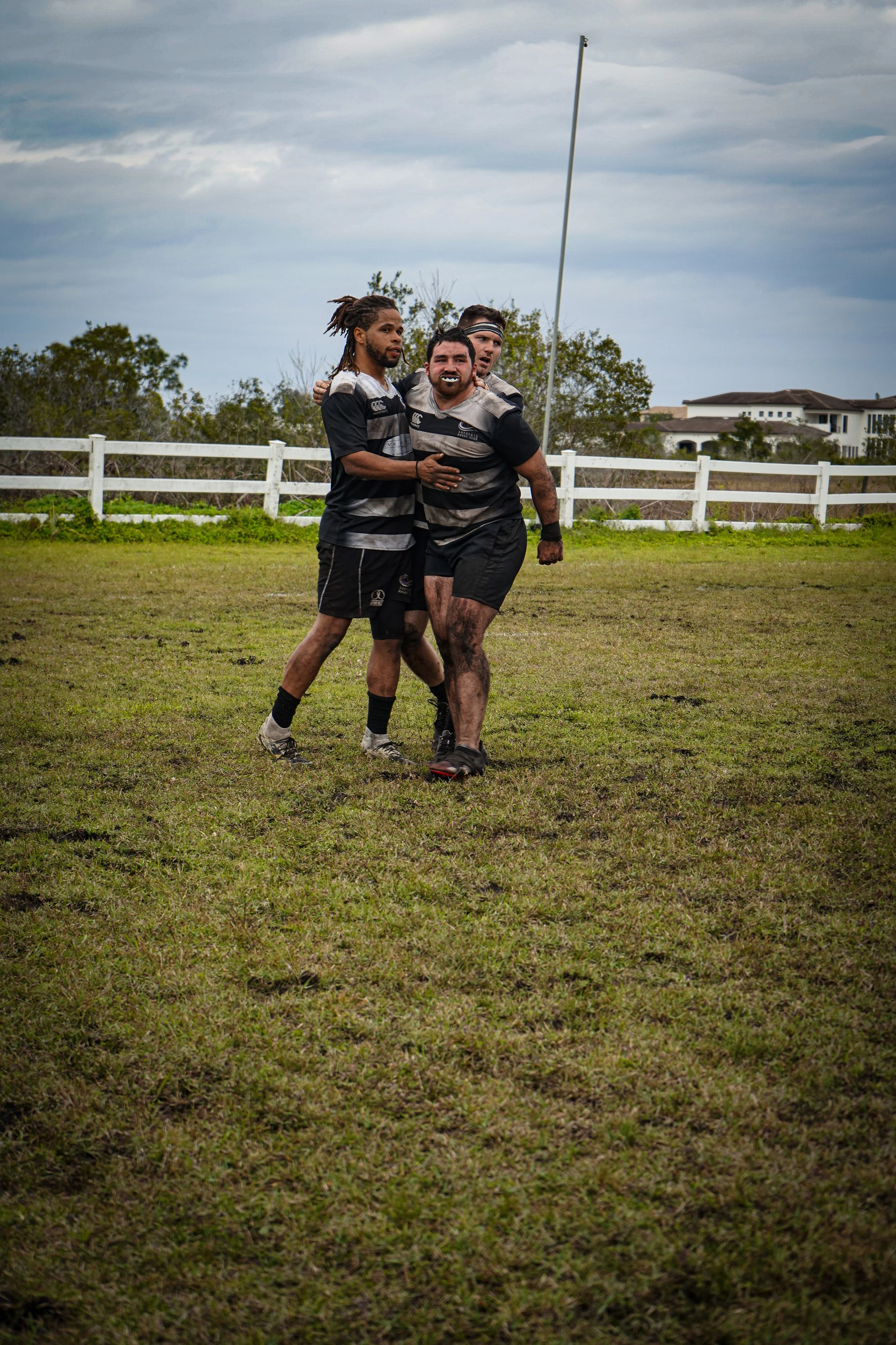 Rugby players celebrating on a muddy field, dressed in black jerseys with gray and white stripes, under a cloudy sky.