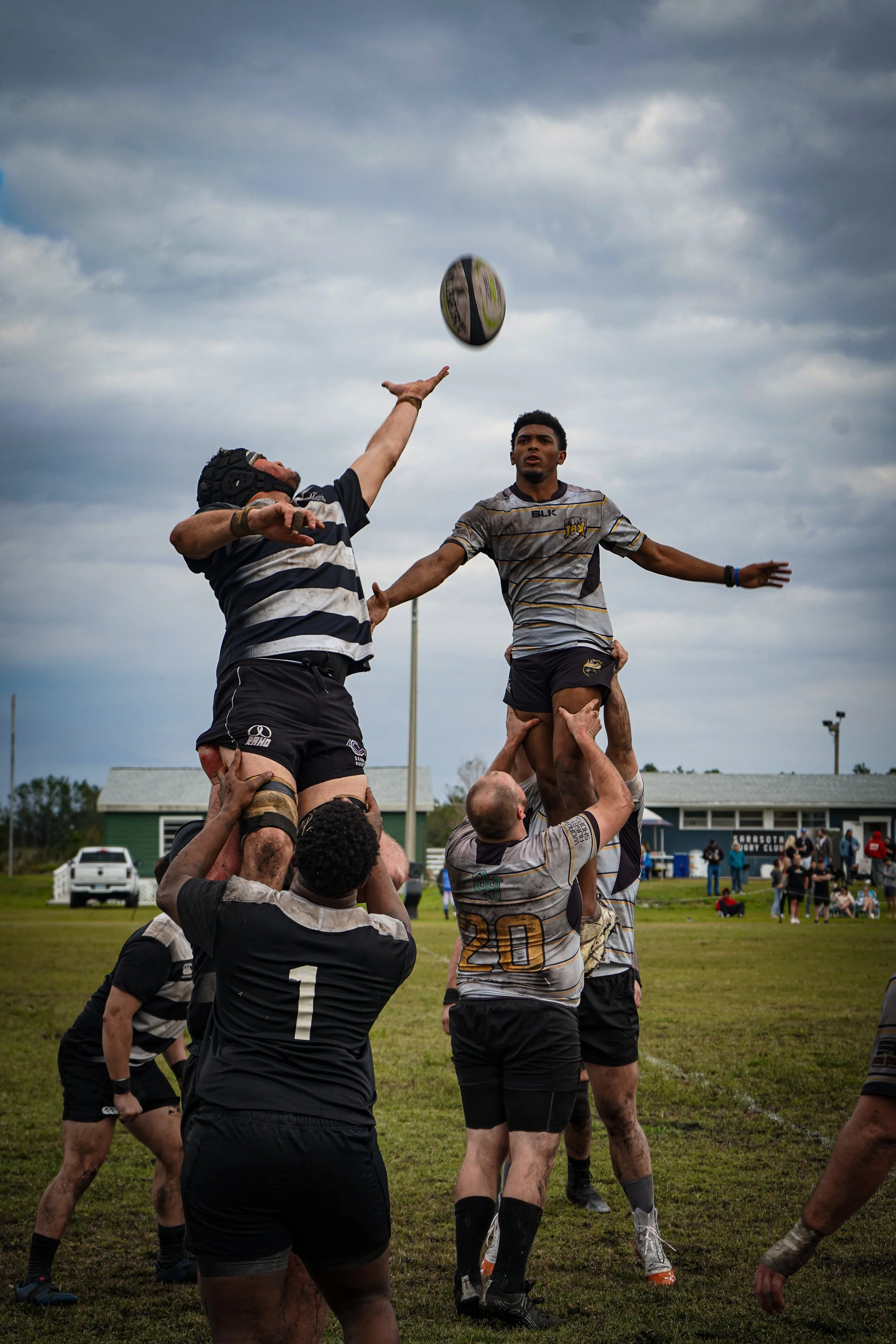Rugby players in a lineout contest on the field, with one player catching the ball as others lift him, under a cloudy sky.