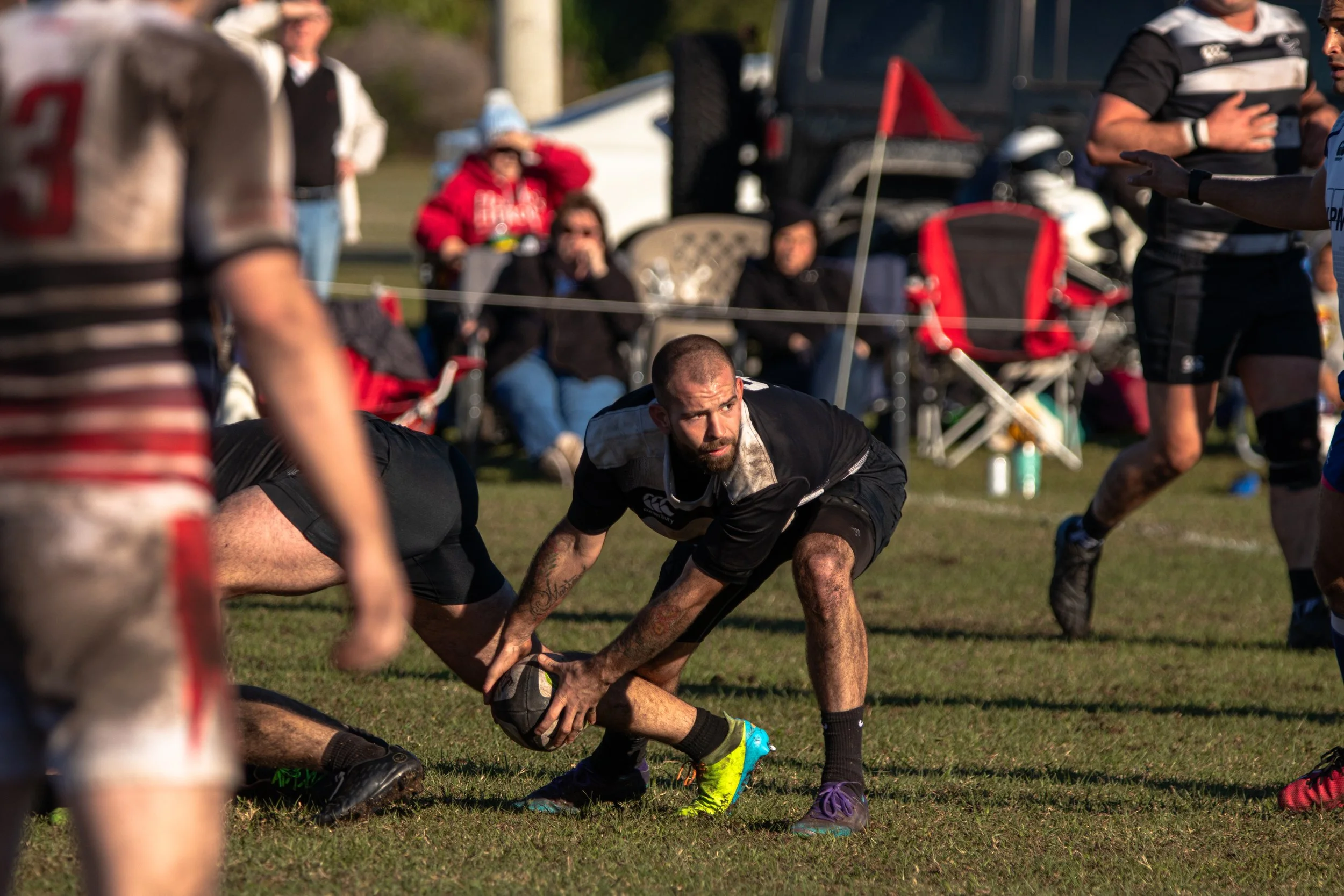 Rugby players in action during a match on a grassy field, with some spectators seated and standing in the background.