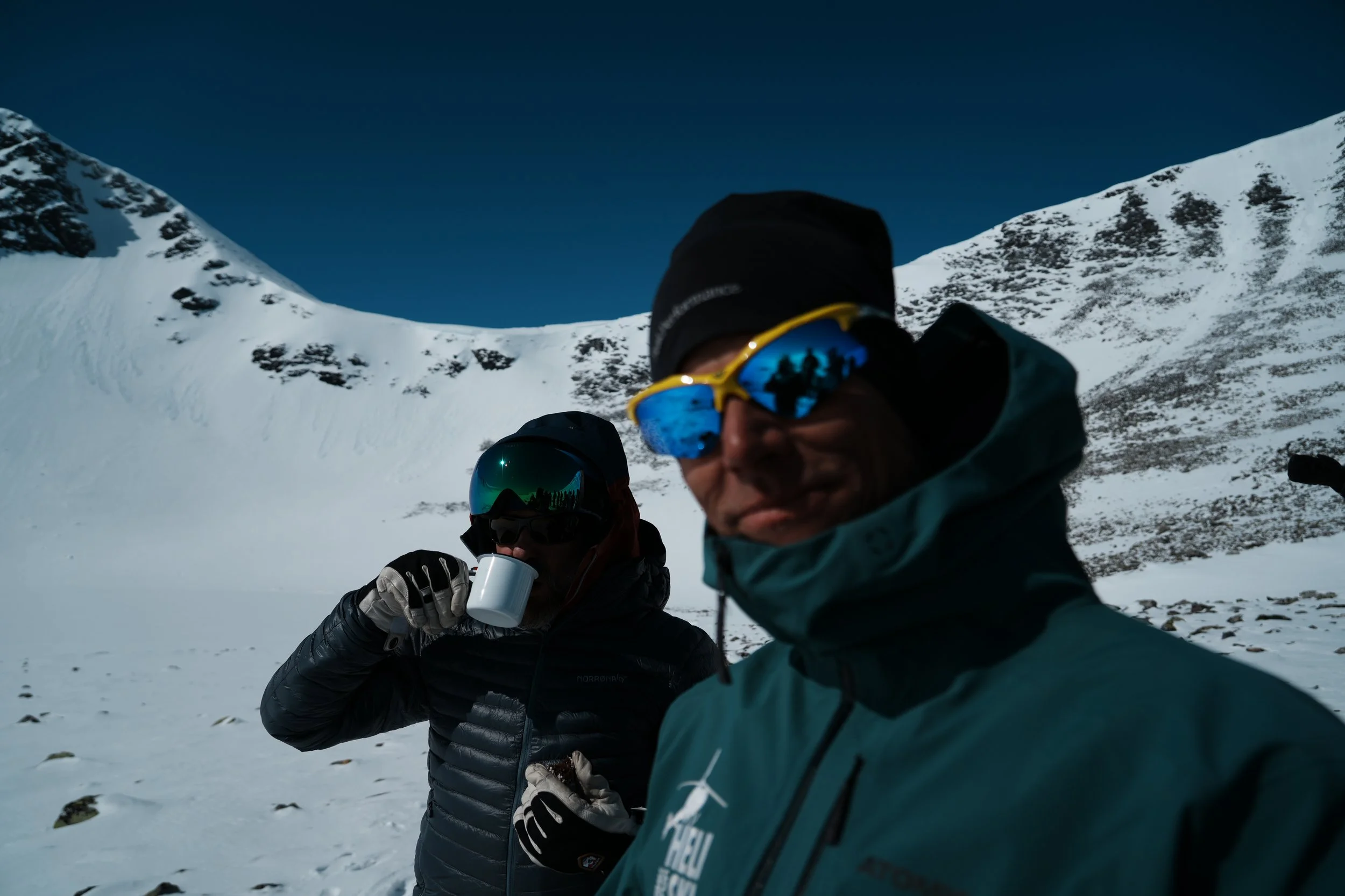 Två personer i vinterkläder och solglasögon som dricker varm dryck i ett snötäckta landskap med snötäckta berg i bakgrunden.