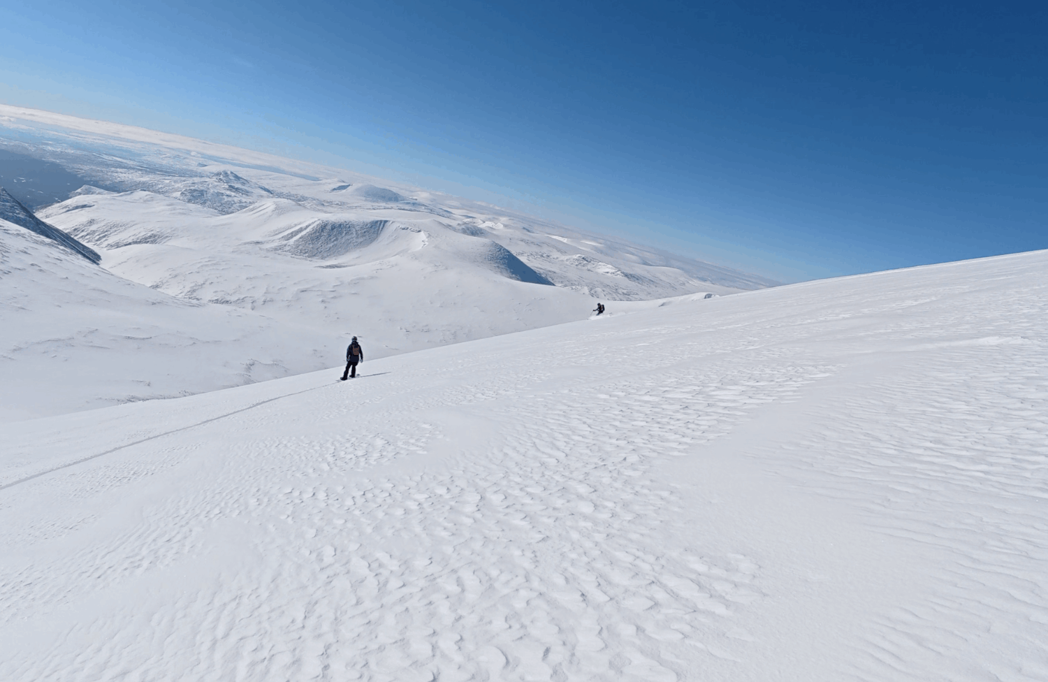 Två personer vandrar i snötäckta fjäll i klart väder med blå himmel, omgivna av snöiga berg.