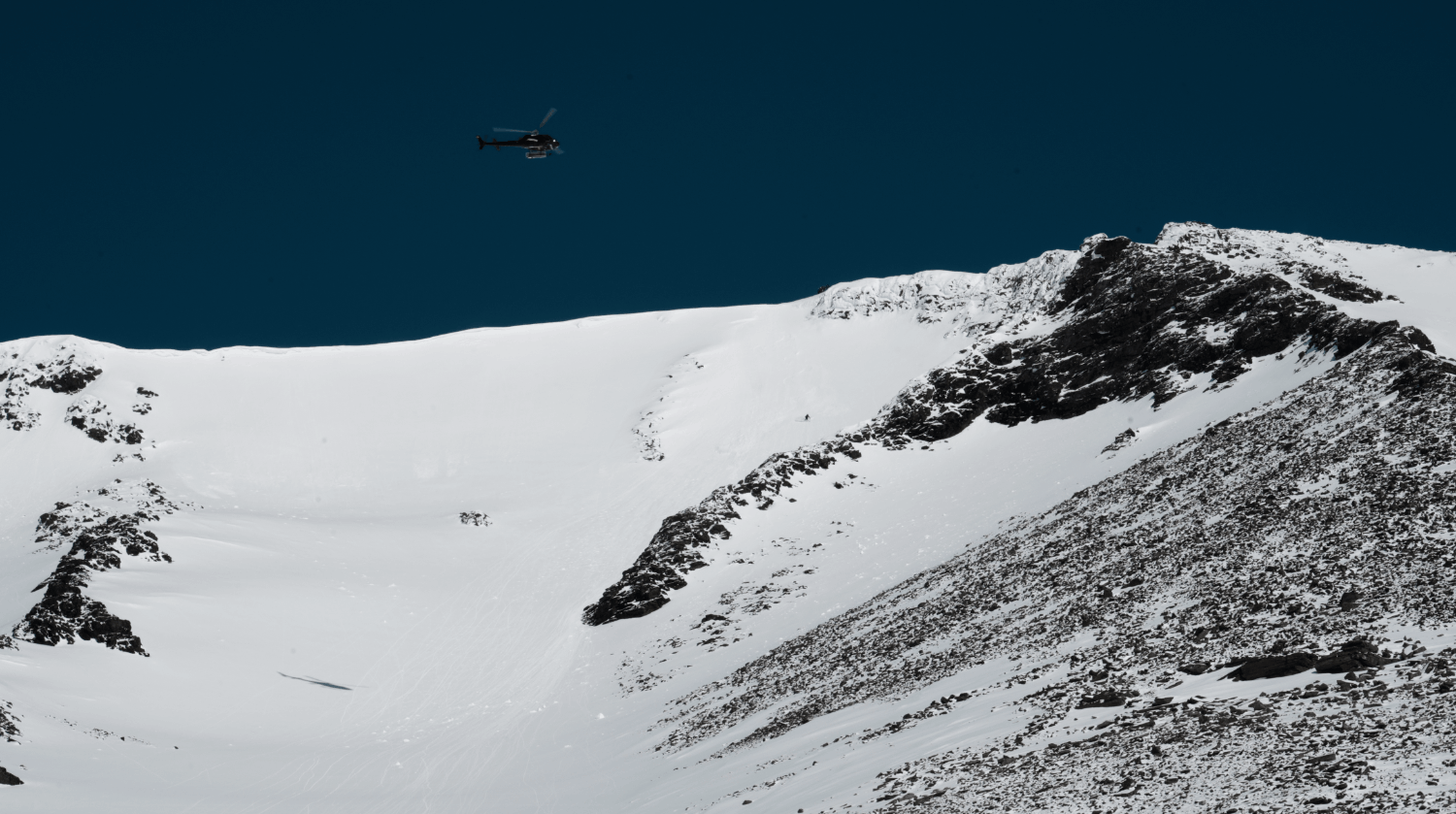 En helikopter flyger över snötäckta berg med en klar blå himmel som bakgrund.