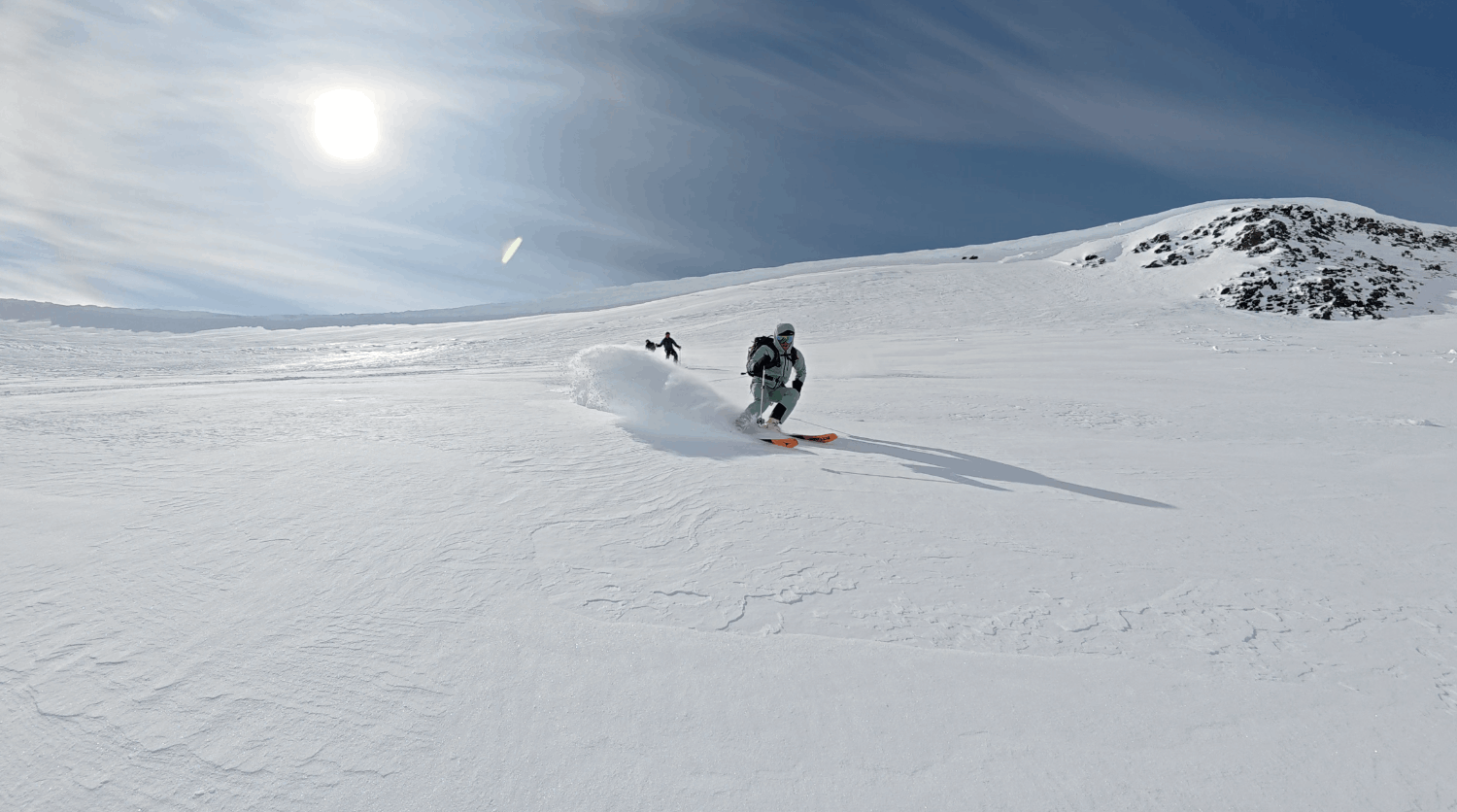 Två skidåkare åker i snön på en bergssluttning under en klar himmel med solen.