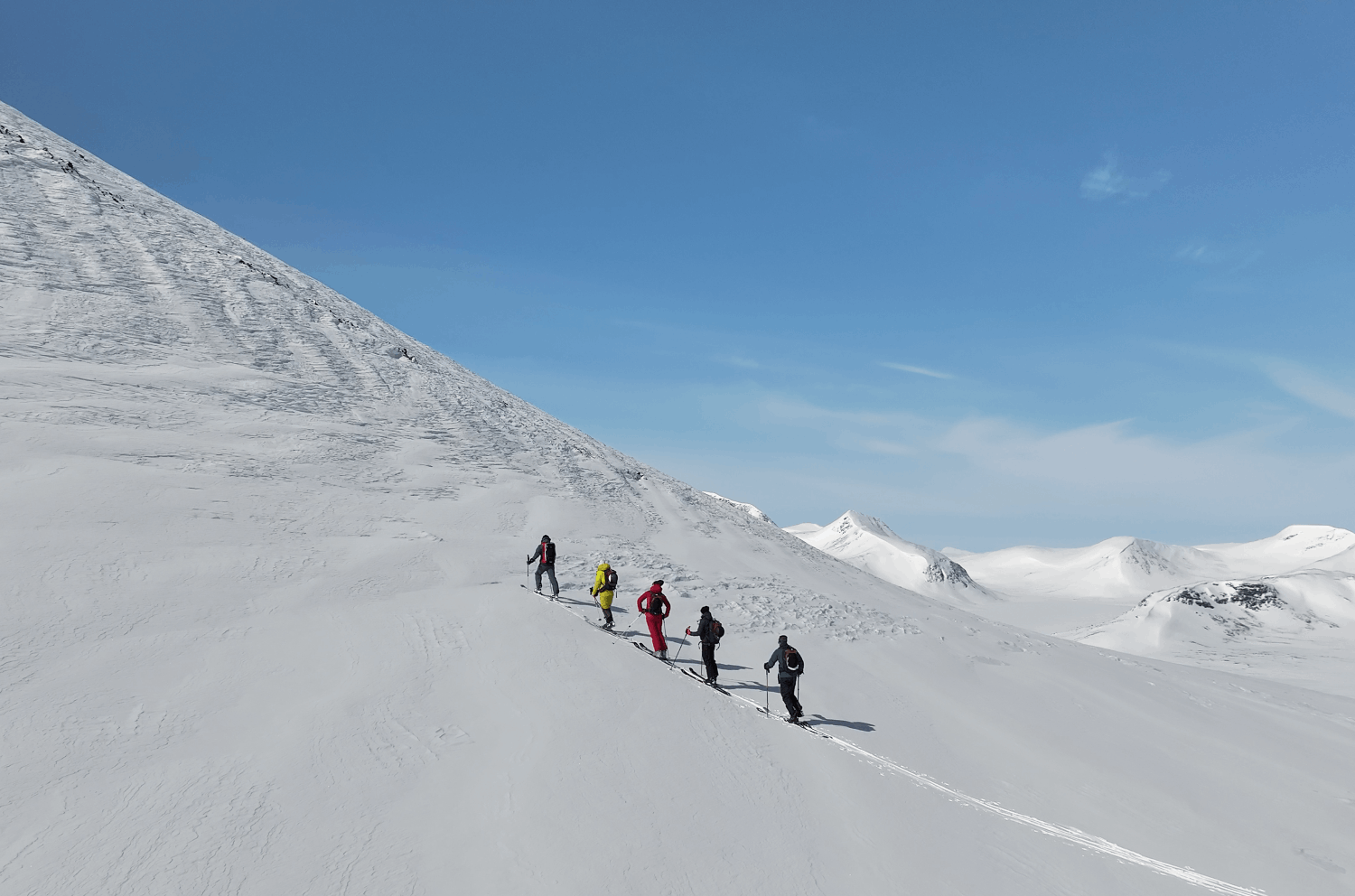 Grupp av fem personer som åker skidor i snötäckta fjällen under en klarblå himmel.