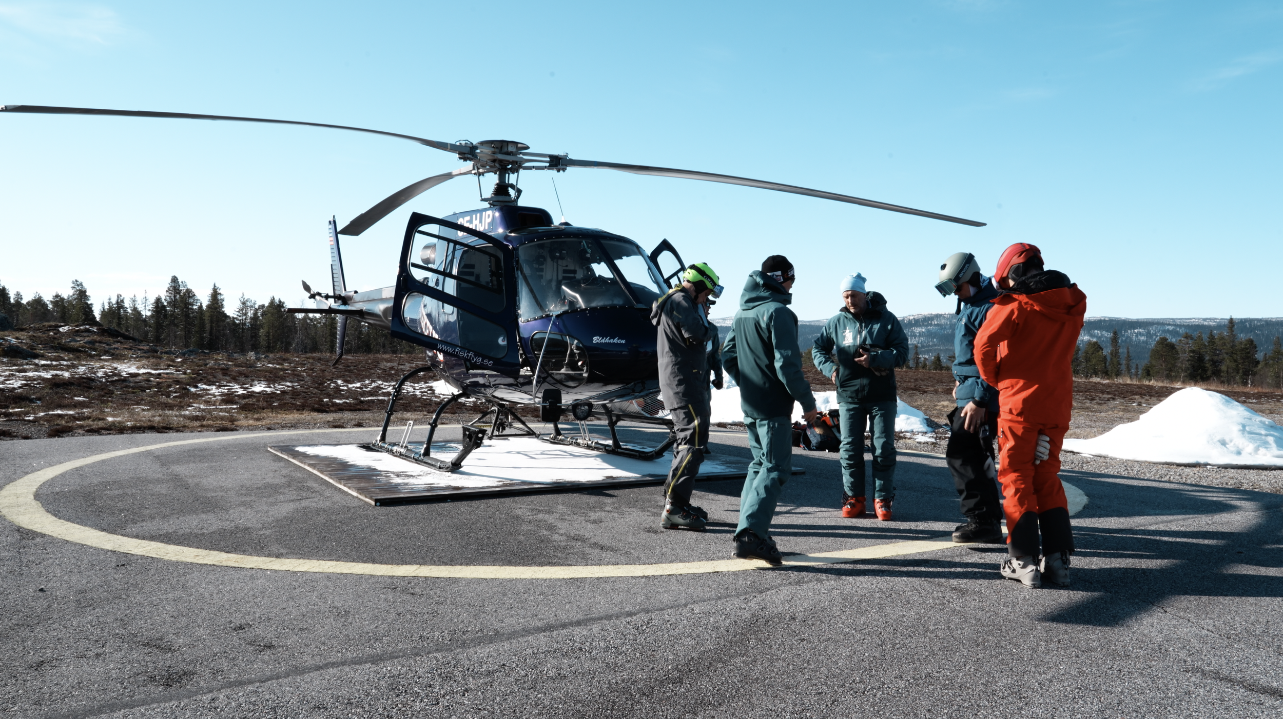 Grupp människor i skidkläder står runt en helikopter på en helikopterplatta i ett vinterlandskap med snö och träd i bakgrunden.