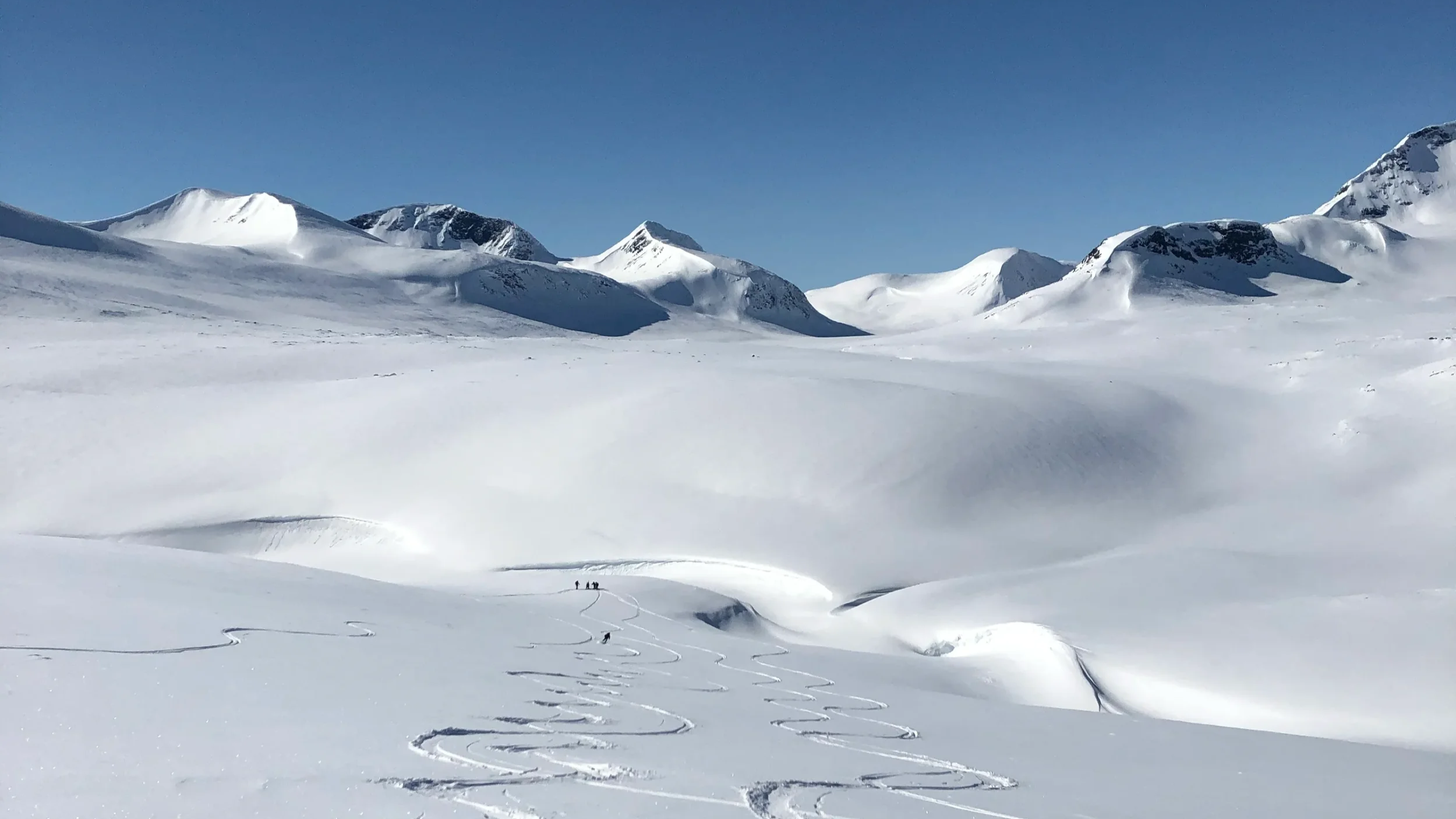 Snötäckta berg och en vidsträckt snöig dal med spår i snön och små silhuetter av personer i fjärran under en klar blå himmel.