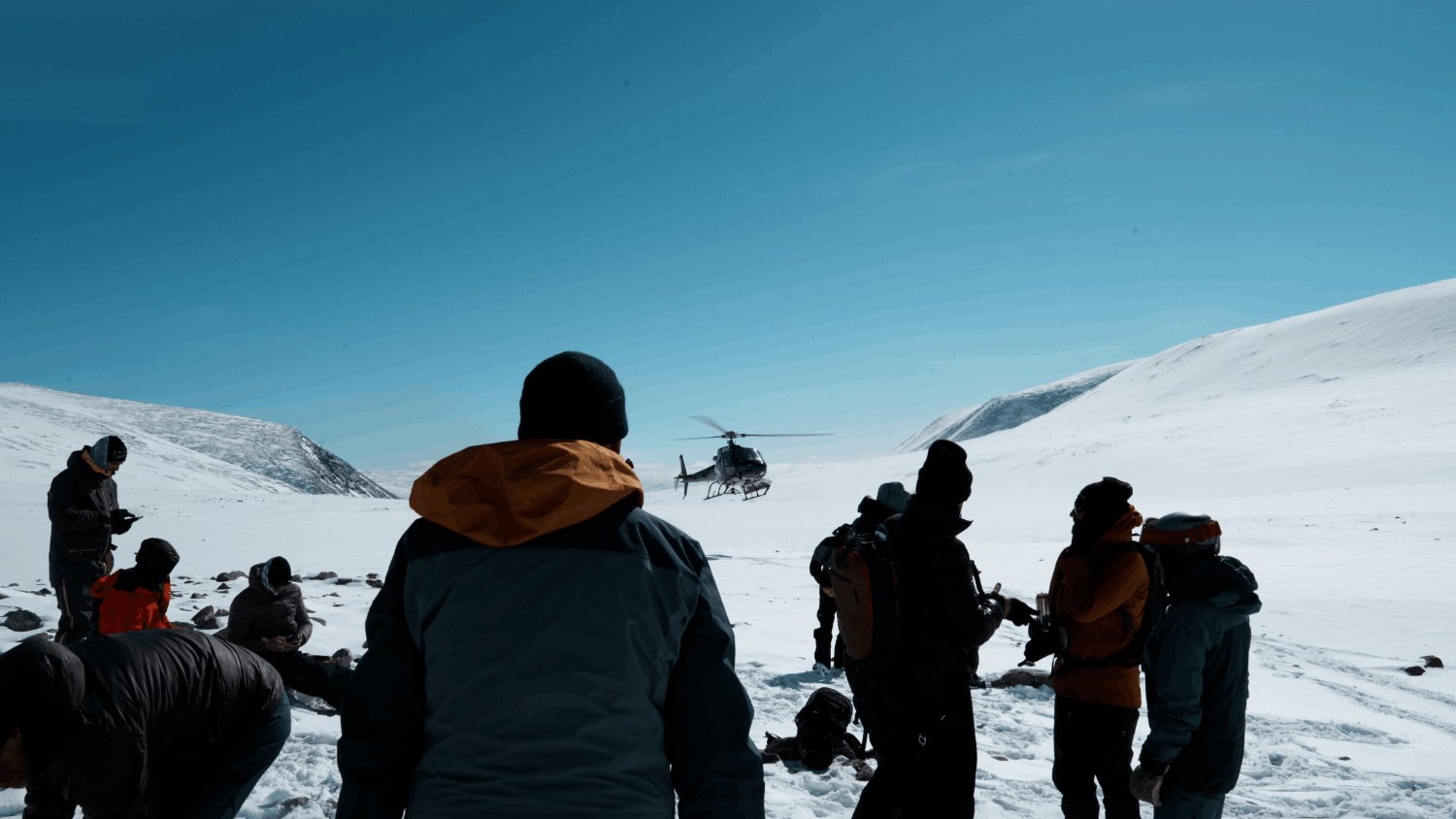 Grupp människor i vinterkläder står på snötäckta landskap medan en helikopter lyfter i bakgrunden.