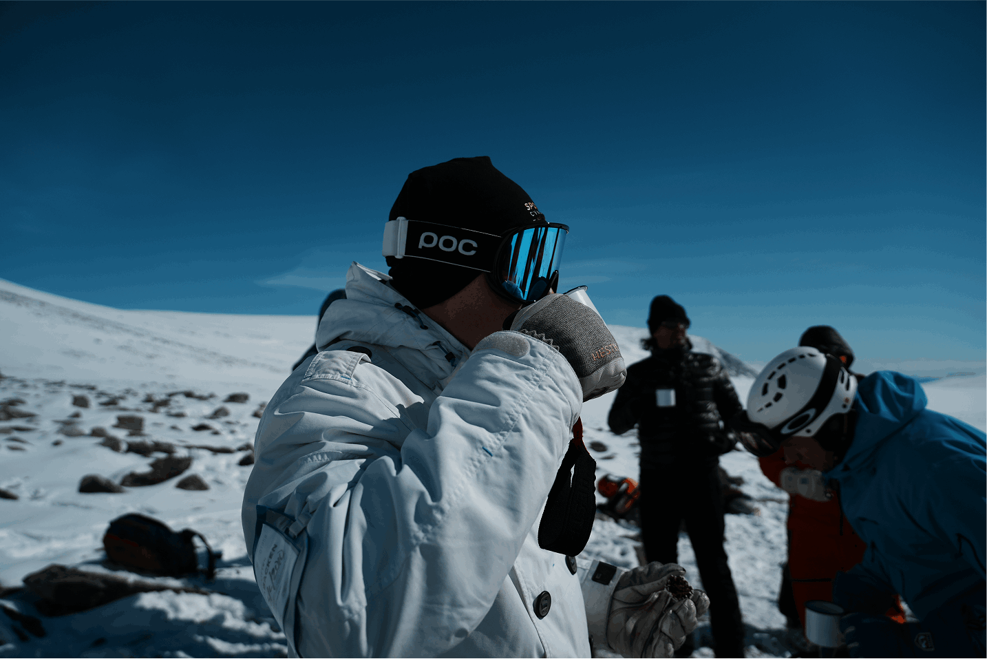 Grupp människor i vinterkläder och skidutrustning på snöig öken i bergsmiljö, med klarblå himmel.