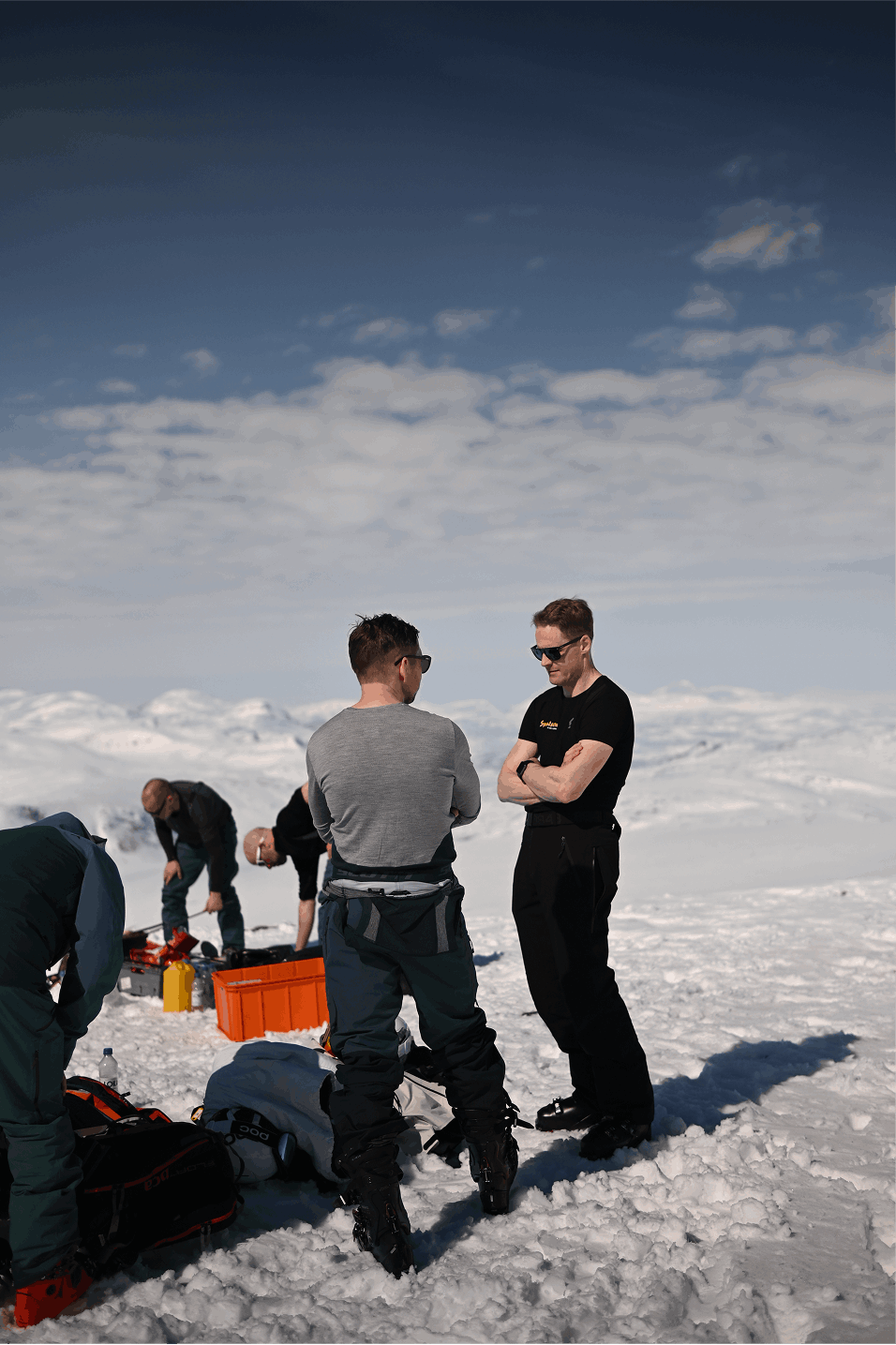 Tre personer står i snön och pratar, medan andra förbereder utrustning runt omkring, i ett snölandskap under en klar himmel.
